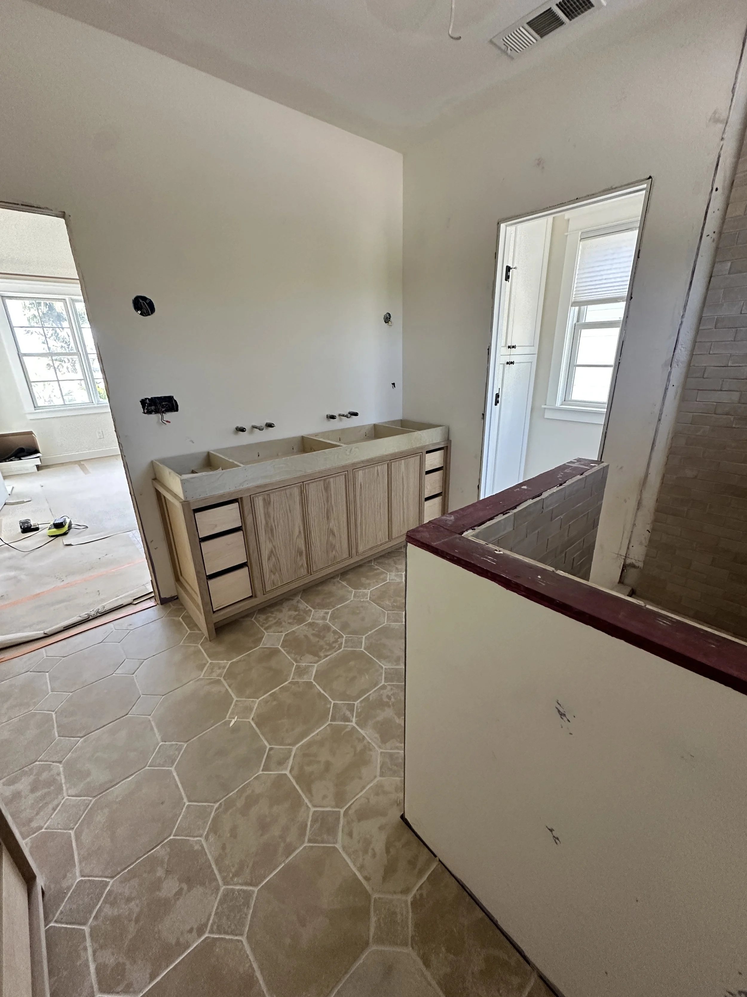 Interior of a house under construction with hexagonal beige floor tiles, unfinished cabinetry, and walls with electrical outlets and exposed wiring.