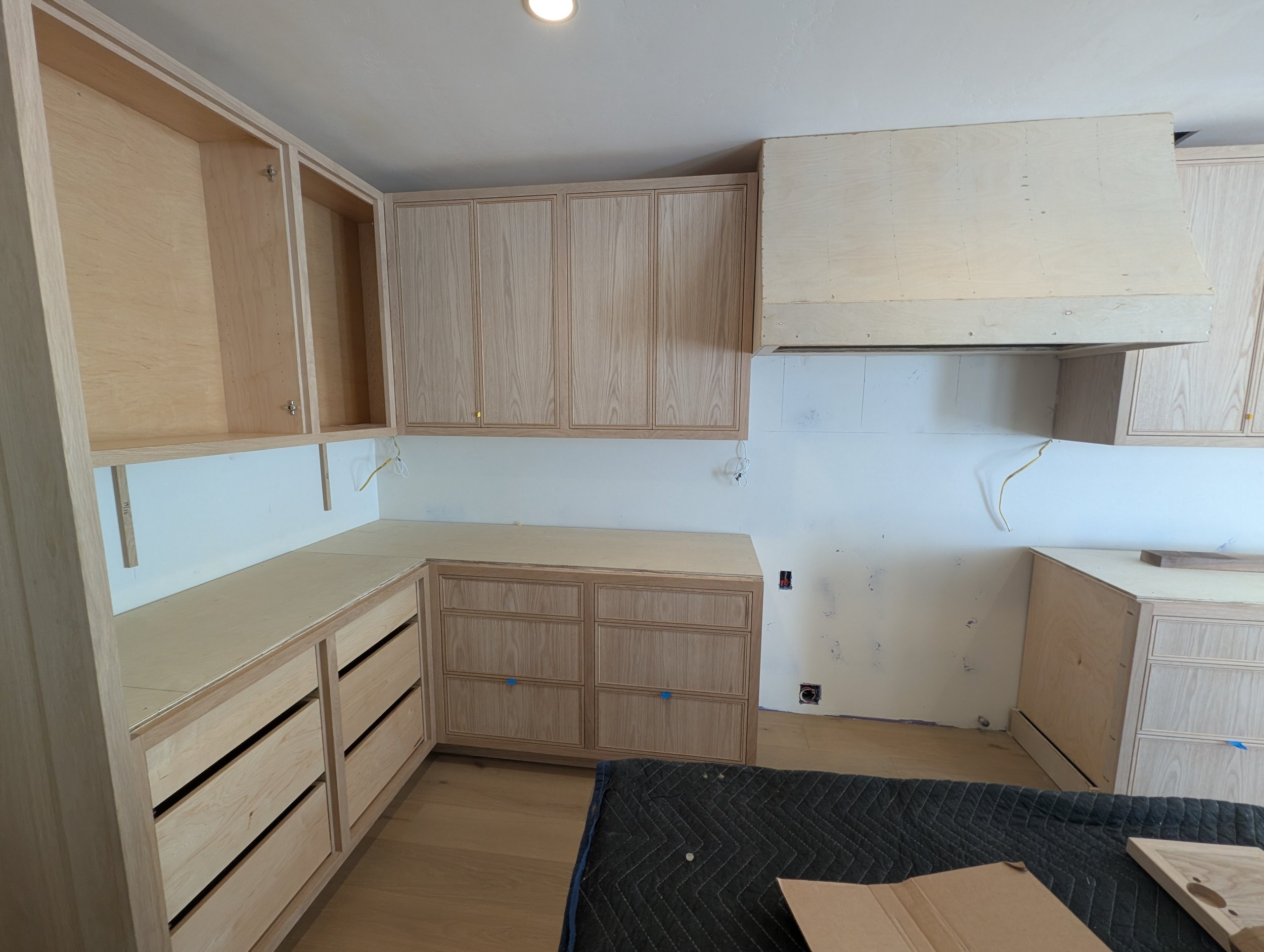 Interior of a room under construction with wooden cabinets and drawers, some of which are not yet finished, and a black quilted mat on the floor.