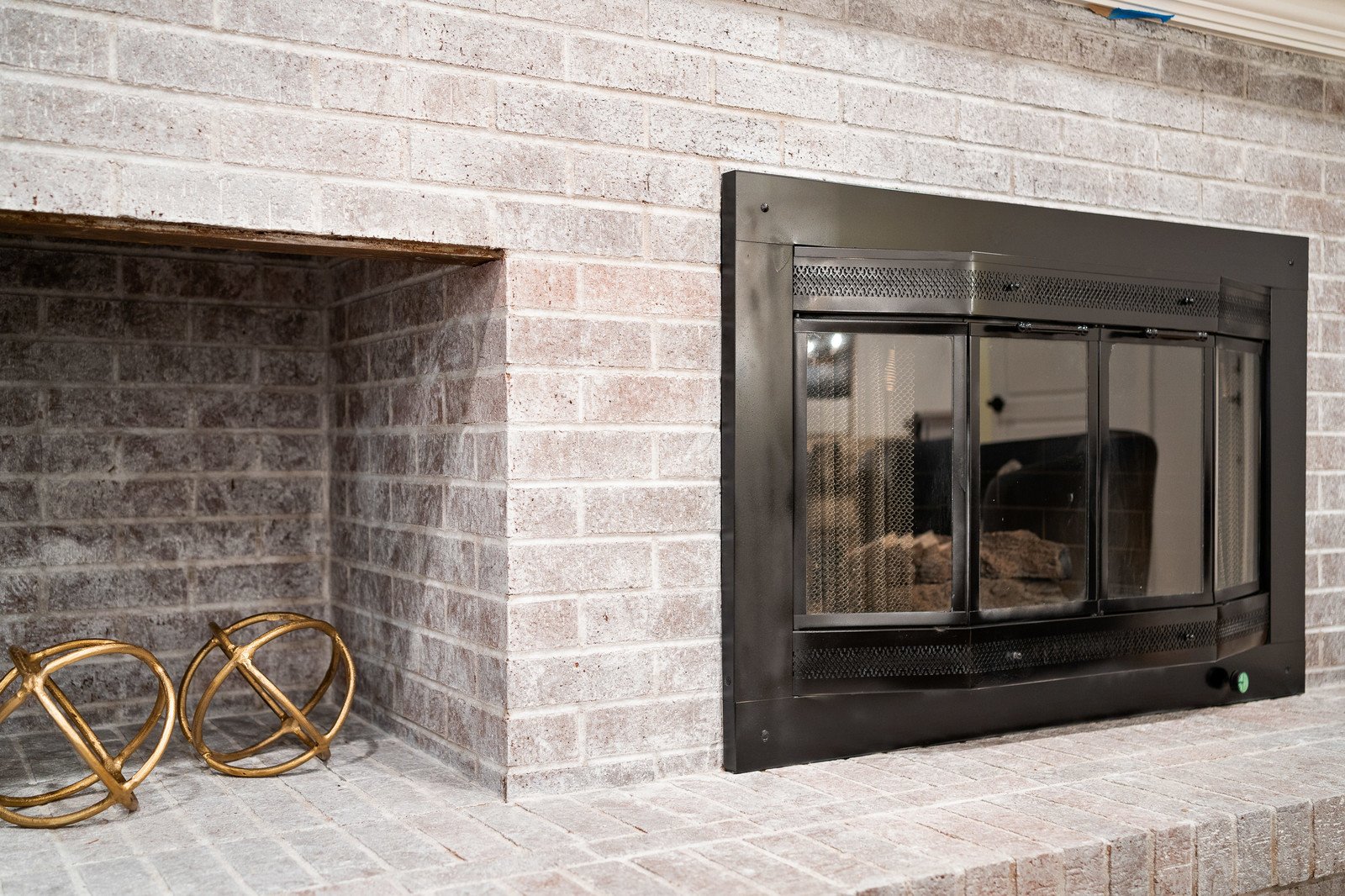 Close-up of a fireplace with a black metal frame and glass doors in a brick wall, with decorative gold fire tools next to it.