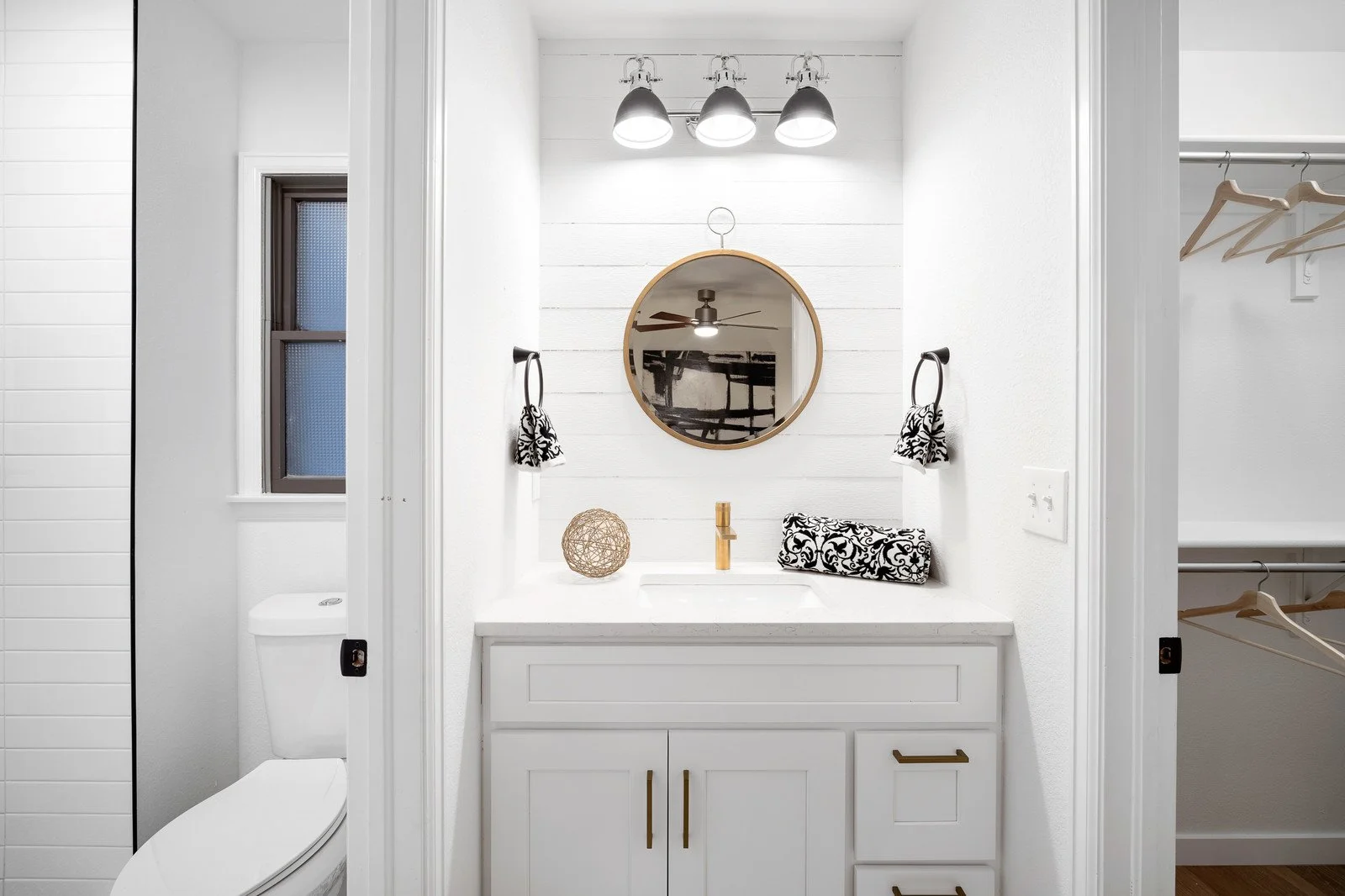A modern bathroom with white walls, a small sink with a white cabinet, a round mirror, a black and white patterned hand towel, a decorative wicker ball, and a gold faucet. There are black wall-mounted towel rings with matching hand towels, overhead l