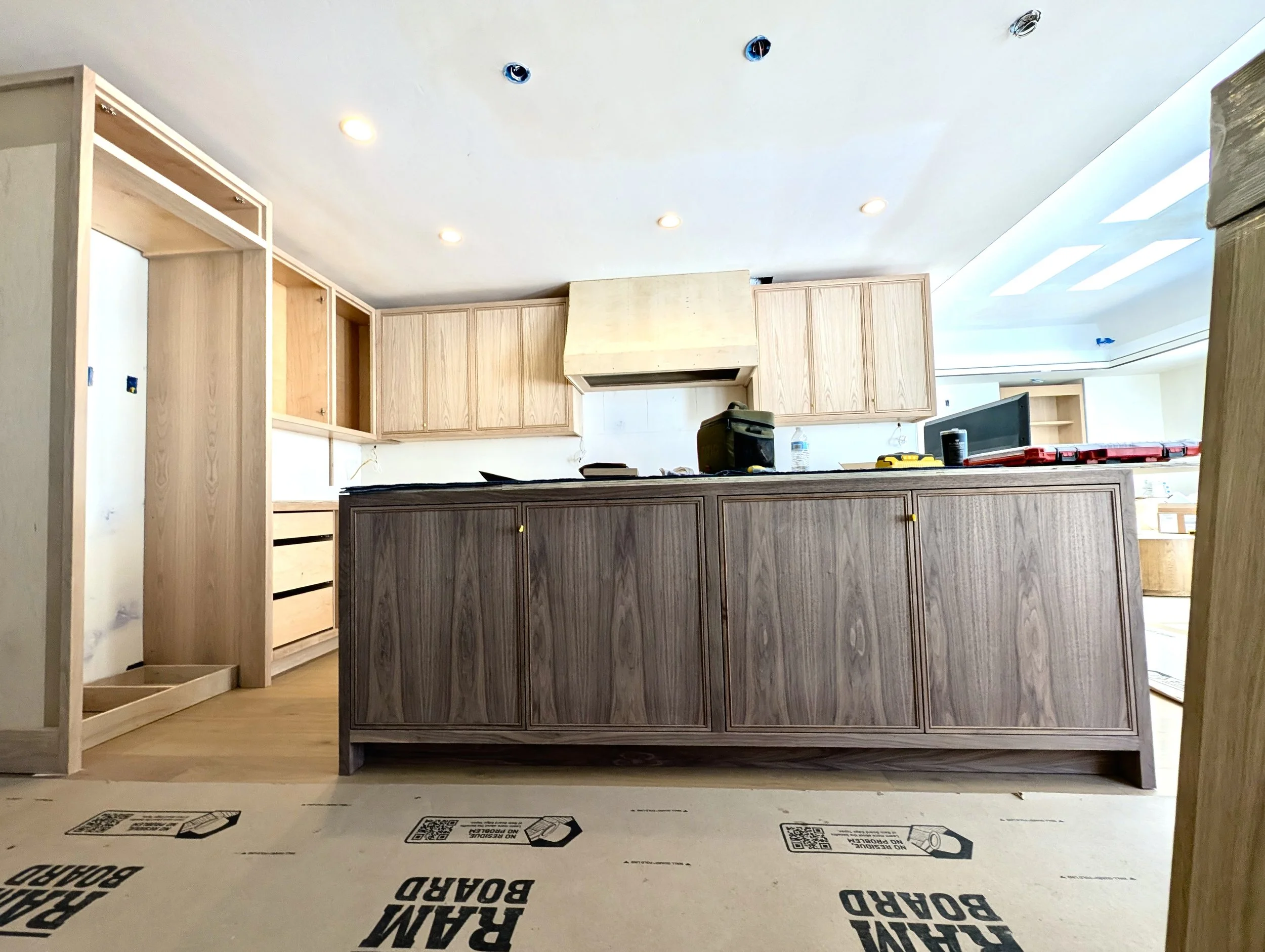 Interior of a house under construction with wooden cabinets, unfinished walls, and flooring with protective paper marked "REAM BOARD" and "DURA-BREST". The ceiling has recessed lighting, and some openings for fixtures or vents. There is construction equipment and materials on the counter.