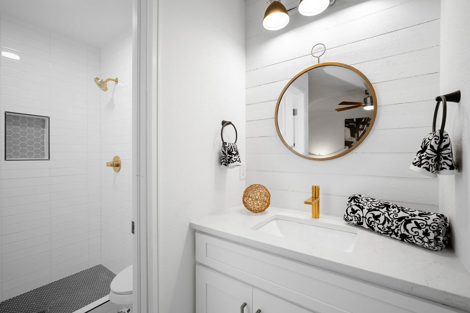 Modern bathroom with white shiplap walls, a round mirror, black and white patterned hand towels, a white vanity with a gold faucet, and a small shower area with white subway tiles and gold fixtures.
