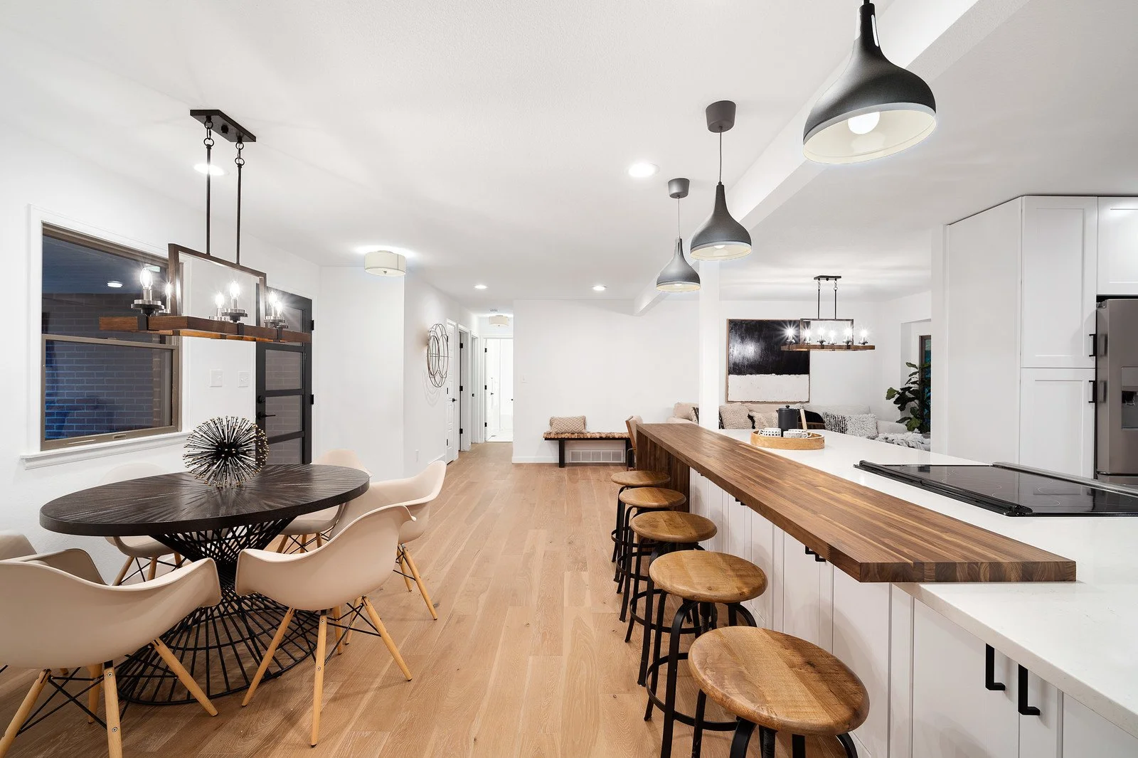 Open-concept kitchen and dining area with white walls, wooden flooring, black pendant lights, a round dining table with beige chairs, and a kitchen island with bar stools.