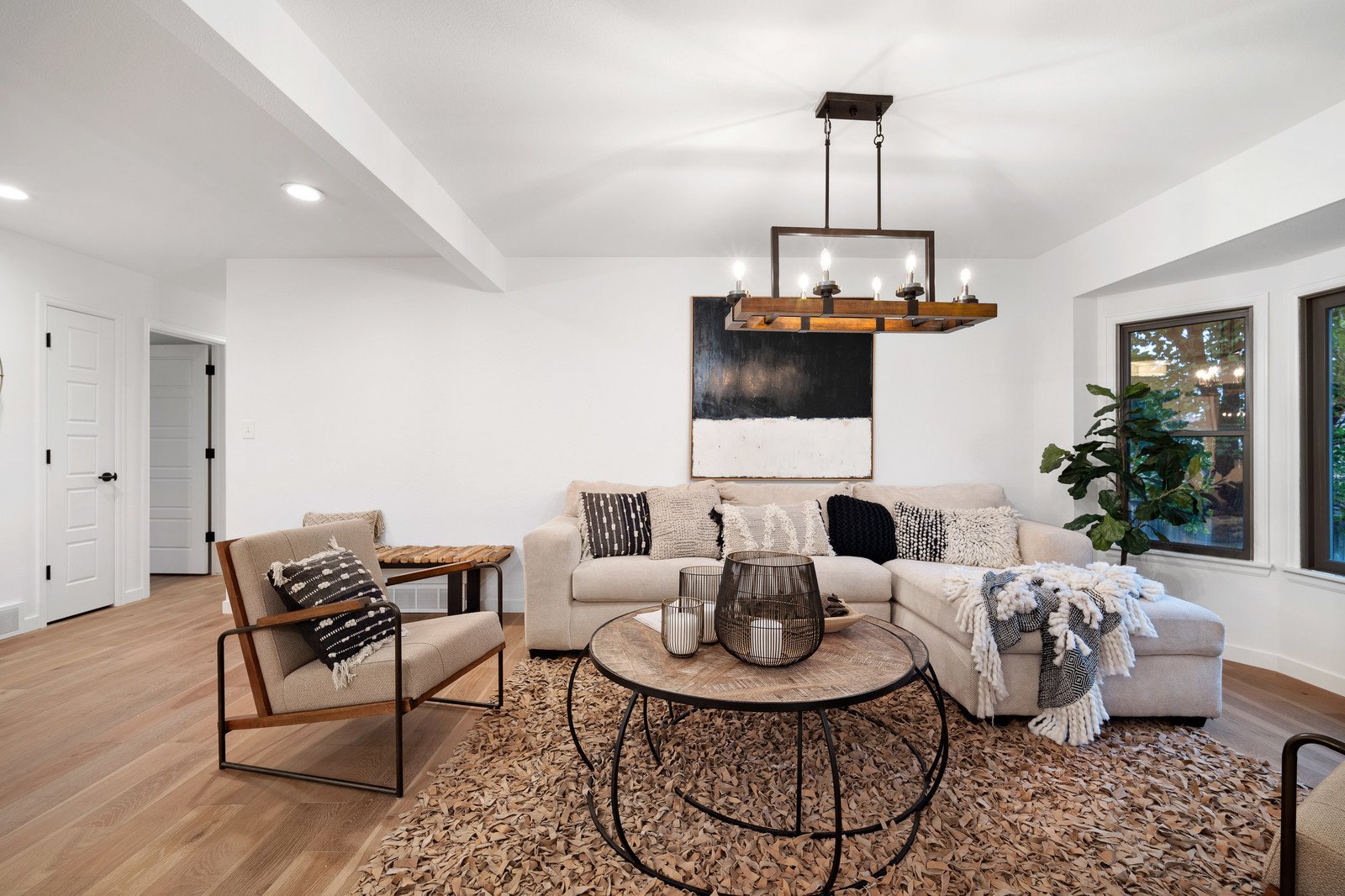 Living room with white walls, beige sectional sofa with black and textured pillows, round wooden coffee table with wireframe decorations, armchair with black and white cushion, large window with greenery outside, potted plant, black abstract painting