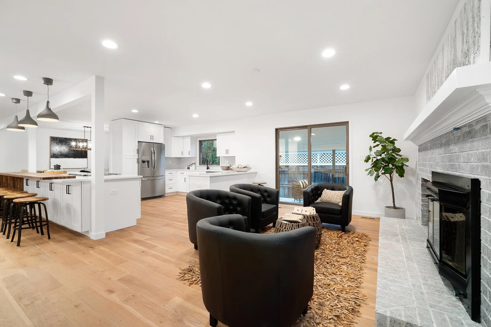 Open-concept living room and kitchen with hardwood flooring, black leather chairs, fireplace, white cabinets, stainless steel refrigerator, and sliding glass door to patio.