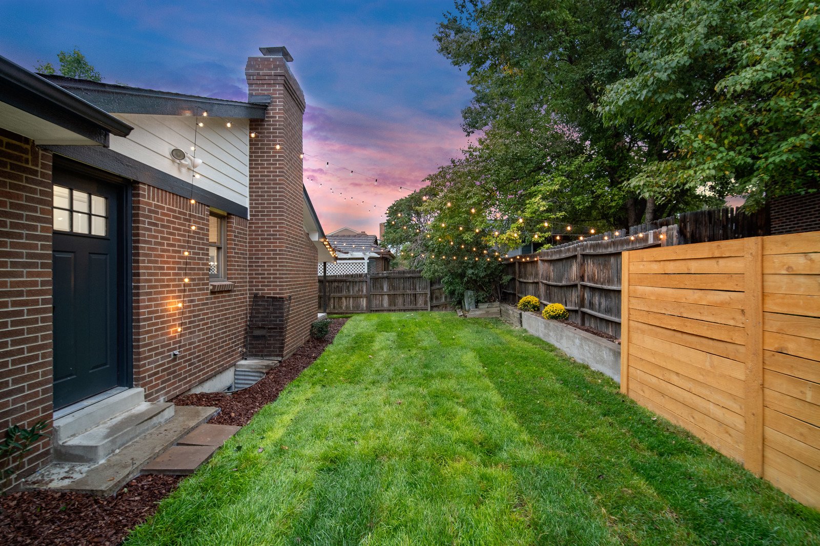 A backyard during sunset with a brick house on the left, string lights hanging across the yard, a grassy lawn, and a wooden fence on the right, with a large tree and colorful sky in the background.