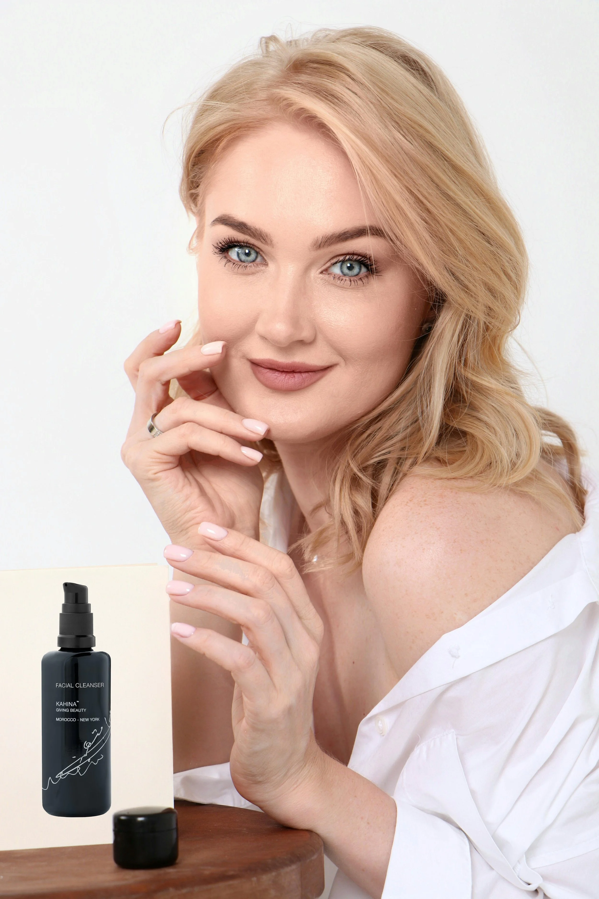 A woman with blonde hair and blue eyes smiling and resting her chin on her hand, with a bottle of facial cleanser on a wooden surface in front of her.