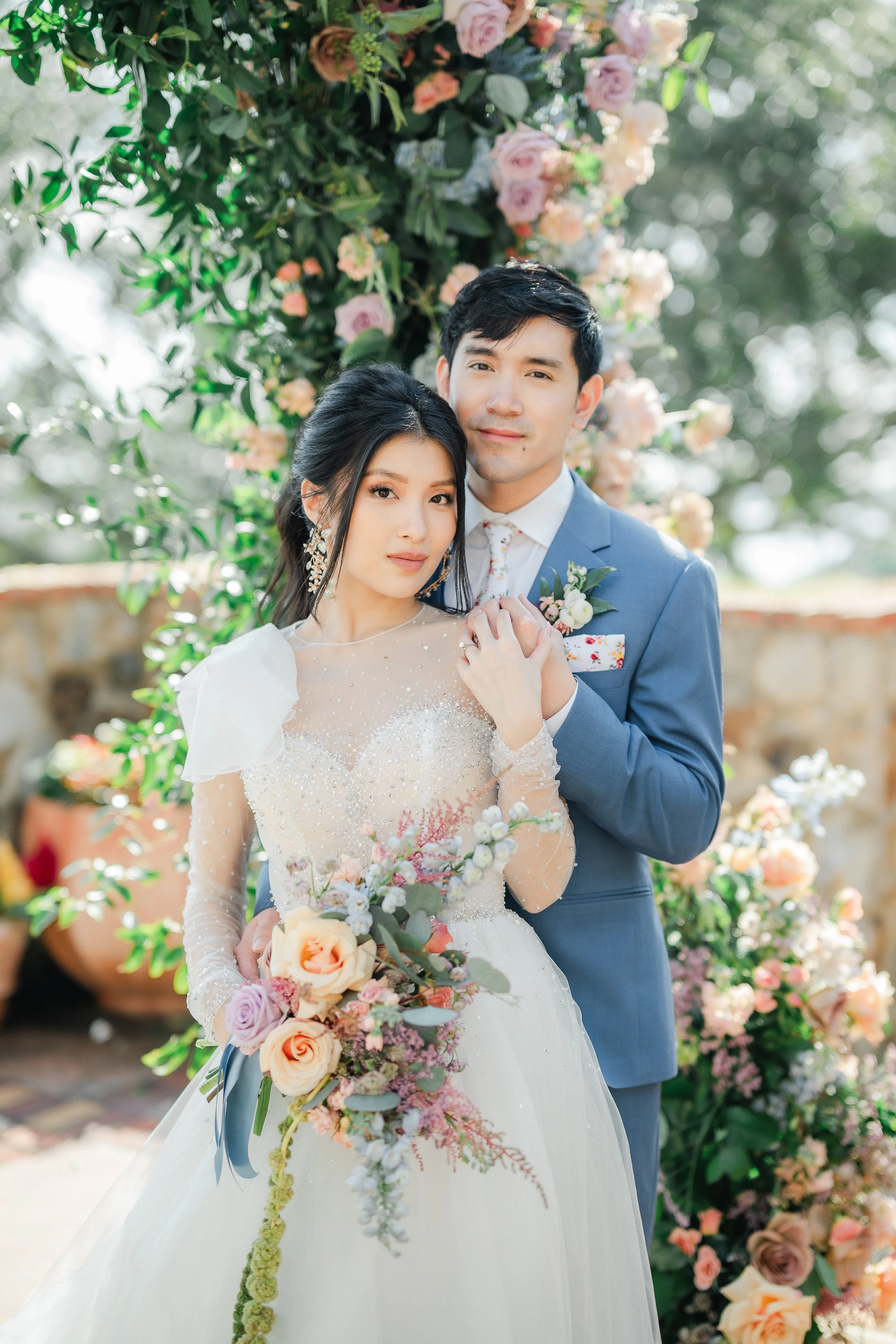 A bride and groom standing together outdoors, surrounded by floral arrangements, with the bride holding a bouquet of pastel roses, peonies, and greenery, both dressed in elegant wedding attire.