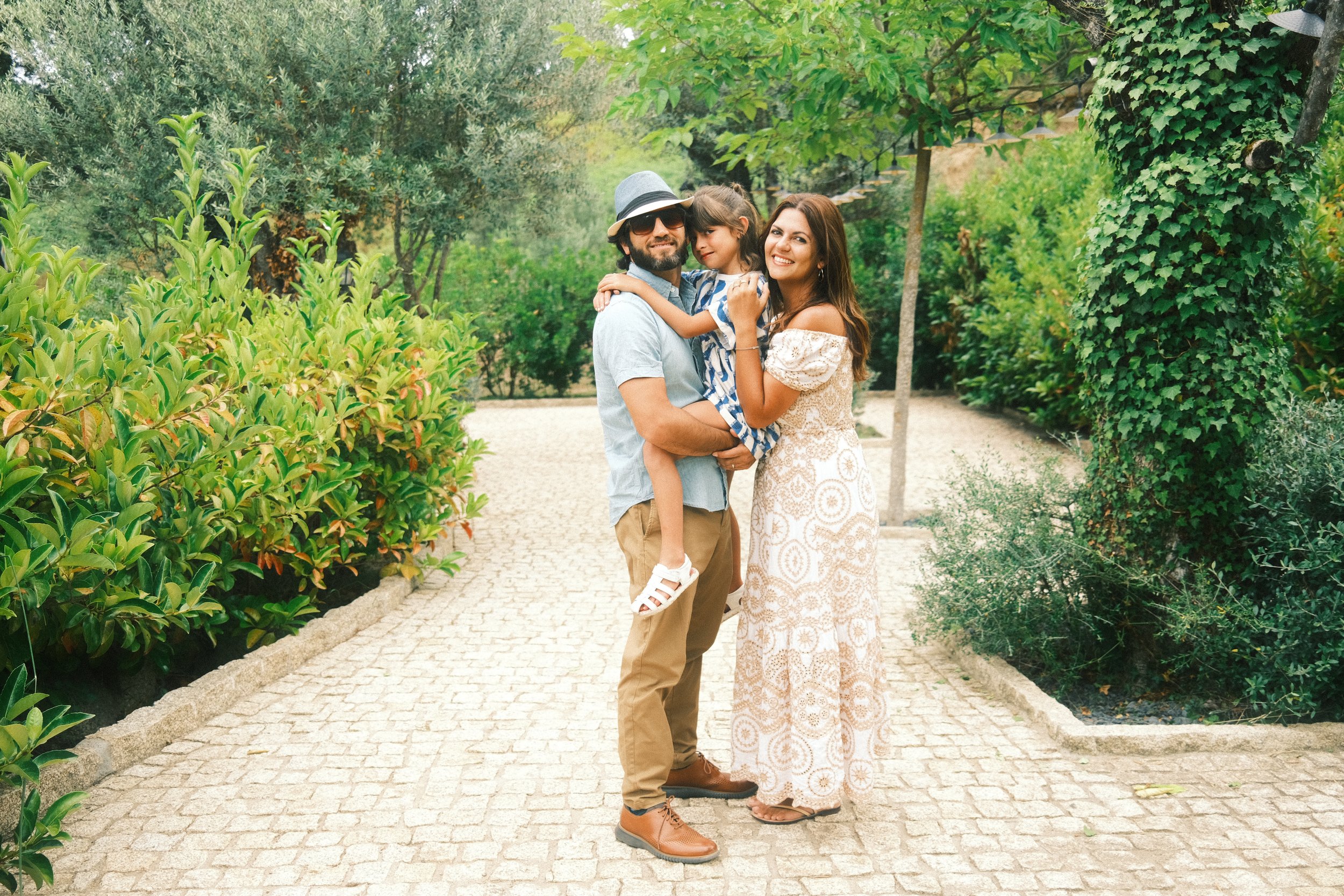 A happy family of three standing on a paved path in a lush park, with greenery and trees around, enjoying a sunny day.