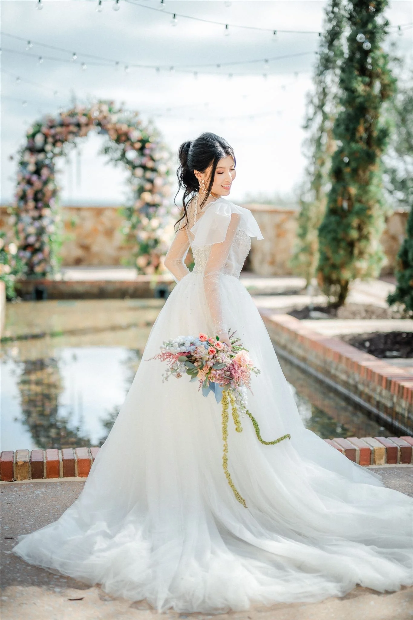 A bride in a white wedding gown holding a bouquet, standing outdoors near a reflective pool, with a floral arch and string lights overhead.