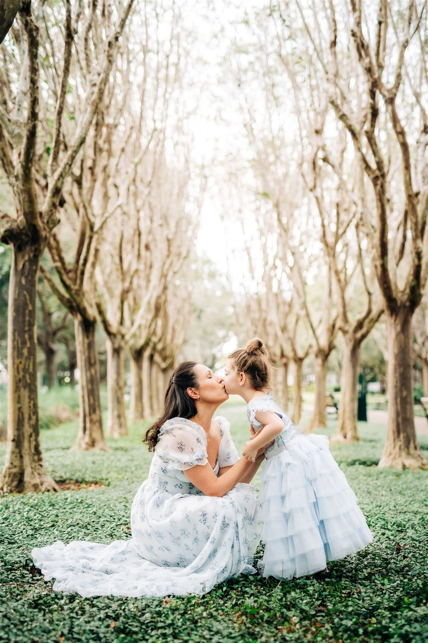 A woman and a young girl are kissing in an outdoor park with a row of leafless trees in the background.