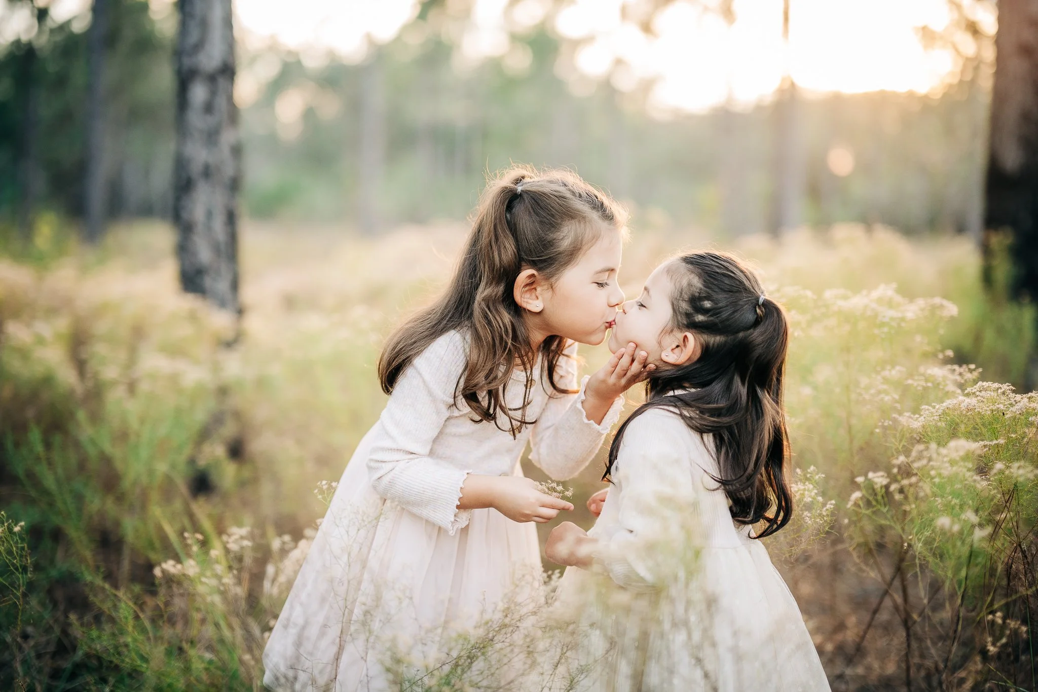 Two young girls in white dresses sharing a kiss in a forest during sunset.