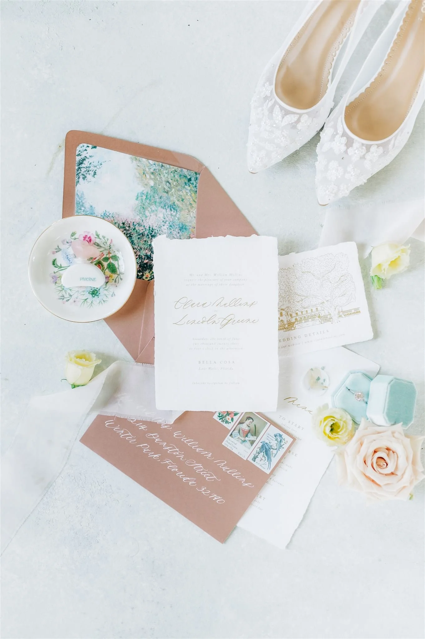 Arrangement of wedding items including white lace high heels, wedding invitations, a ring box, flowers, a postcard, and a jewelry box.