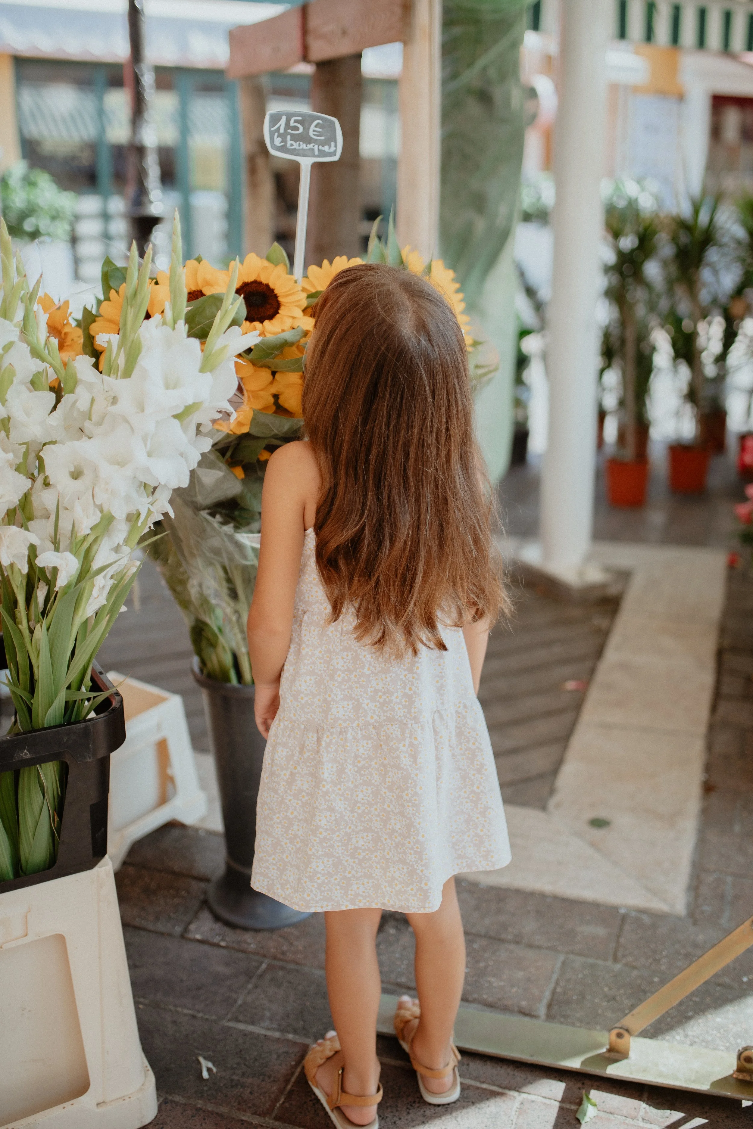A young girl with long brown hair, wearing a white dress with a floral pattern, stands in front of a flower stand at a market, looking at sunflower and white lilies bouquets.