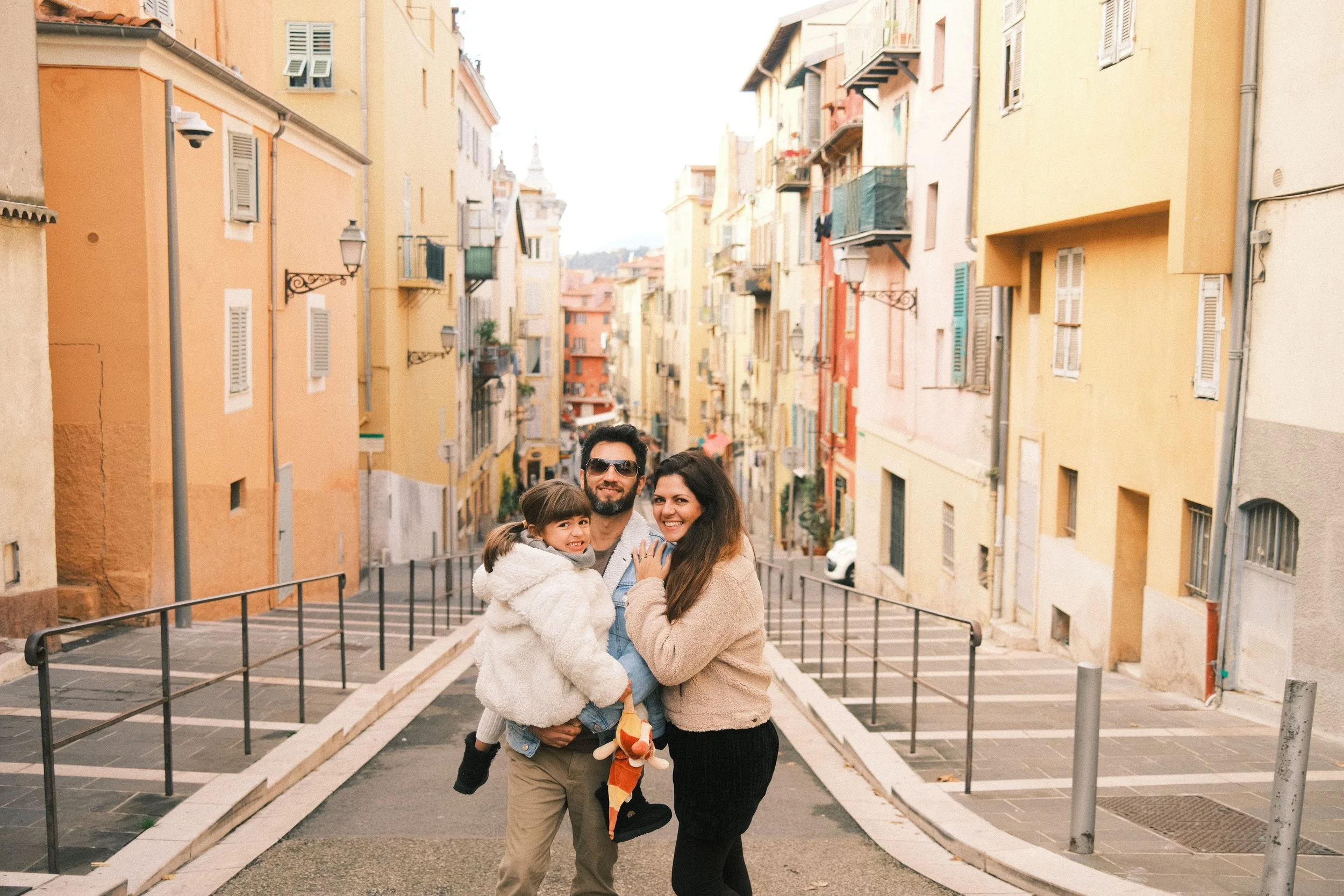 A family of three, a man, a woman, and a girl, smiling and posing together on a street with colorful buildings in the background.