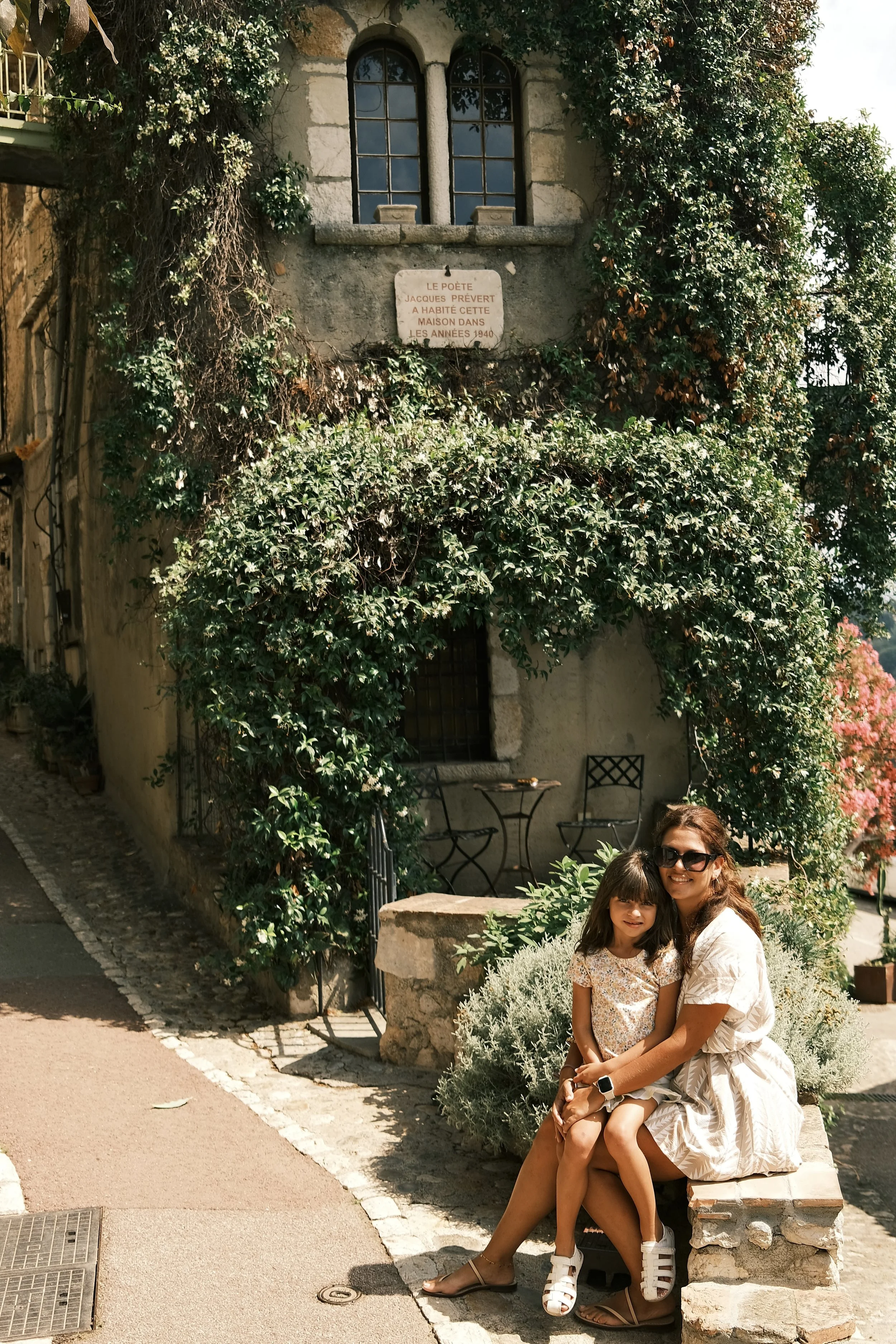 A woman and a young girl sitting on a stone bench in front of a quaint cottage covered in ivy, with a sign above the window that reads in French about the poet Jacques Prévert.