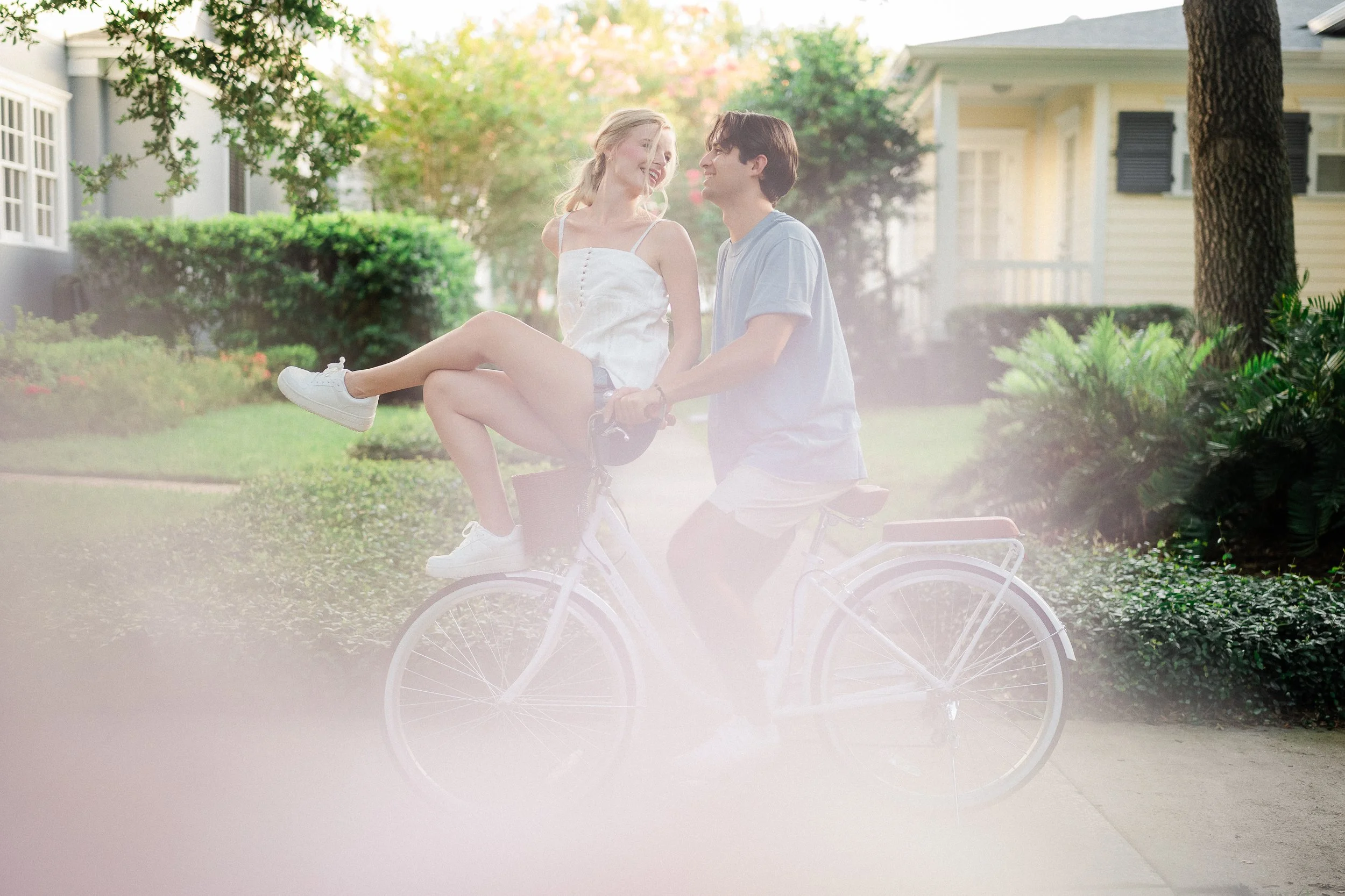 A young couple riding a bicycle outdoors, with the girl sitting on the handlebars and smiling at the guy, in front of a house with greenery.
