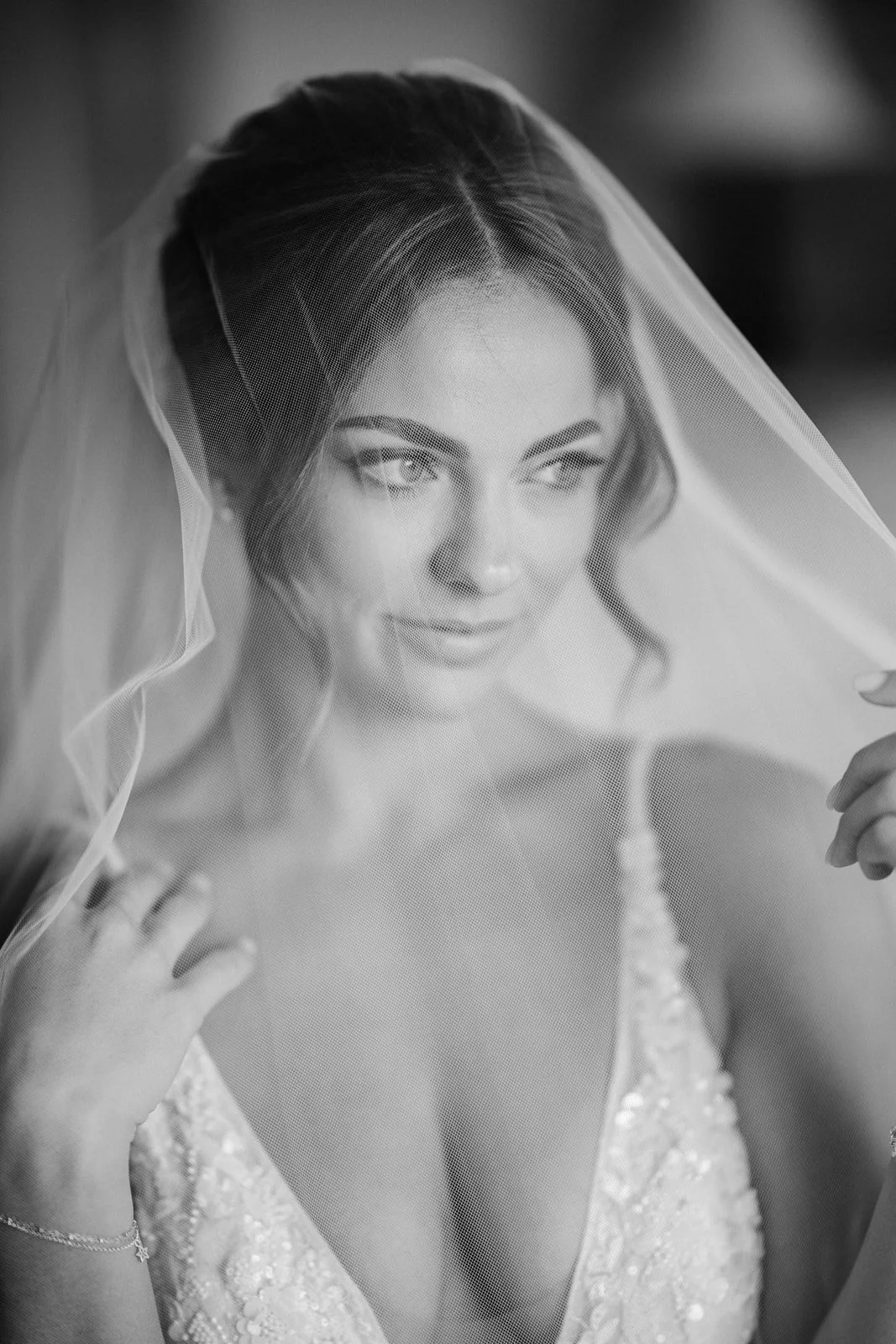 Black and white photo of a bride with a veil, styled hair, and makeup, wearing a dress with lace details.