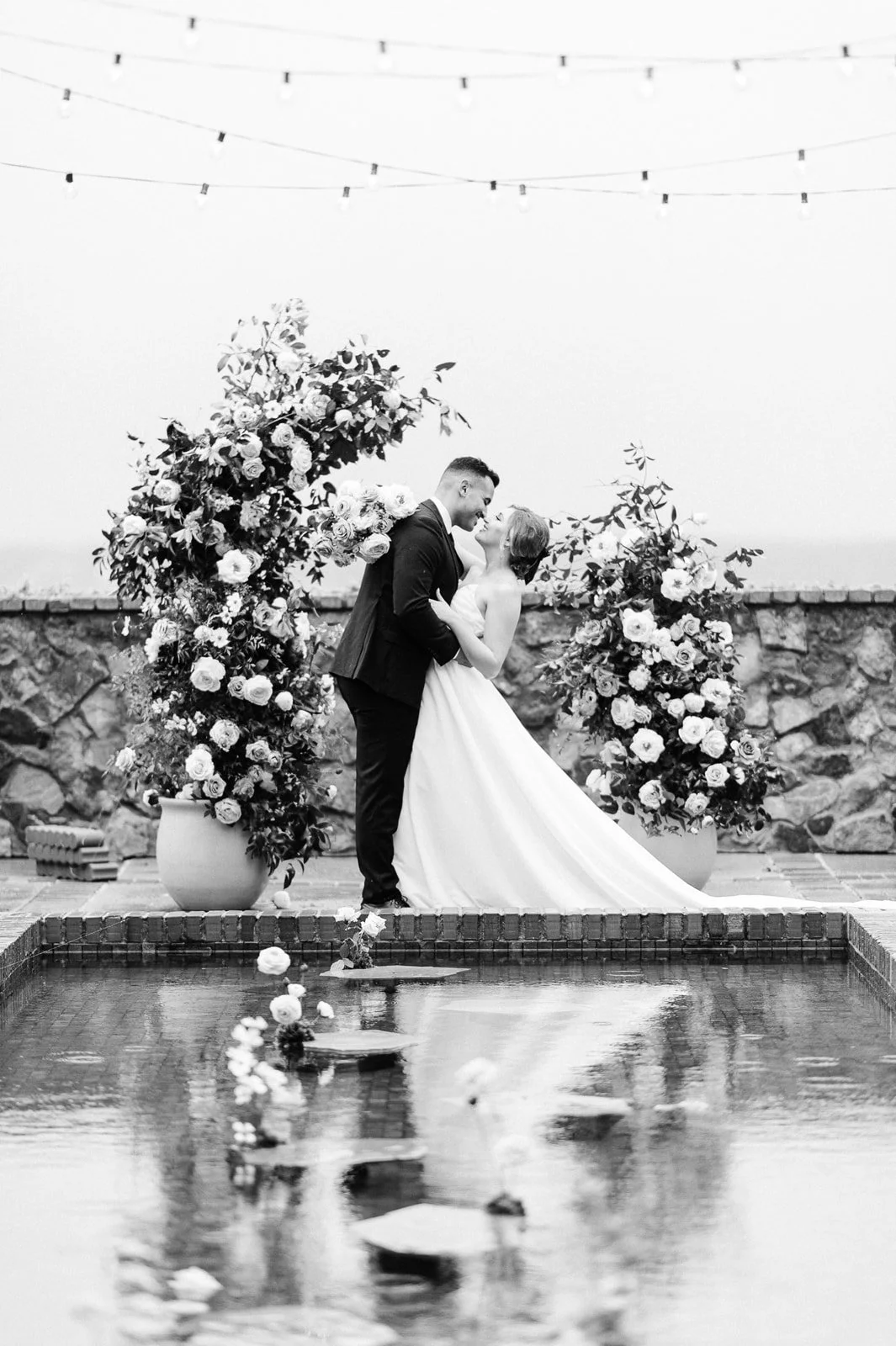A black and white photo of a bride and groom dancing beside a small pool, with large flower arrangements behind them and string lights above, in an outdoor wedding setting.