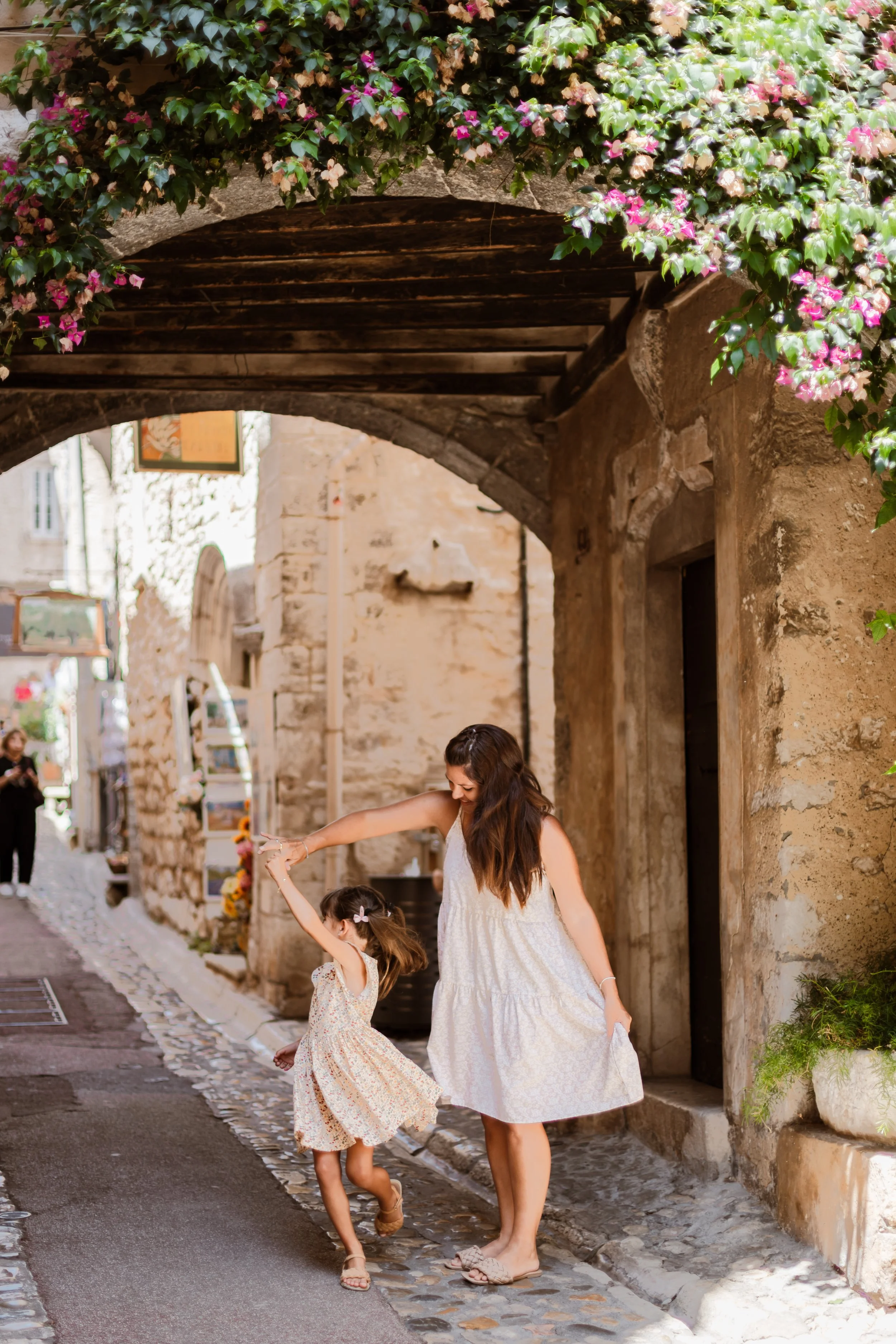 A woman and a young girl dancing and holding hands on a cobblestone street in a historic town.