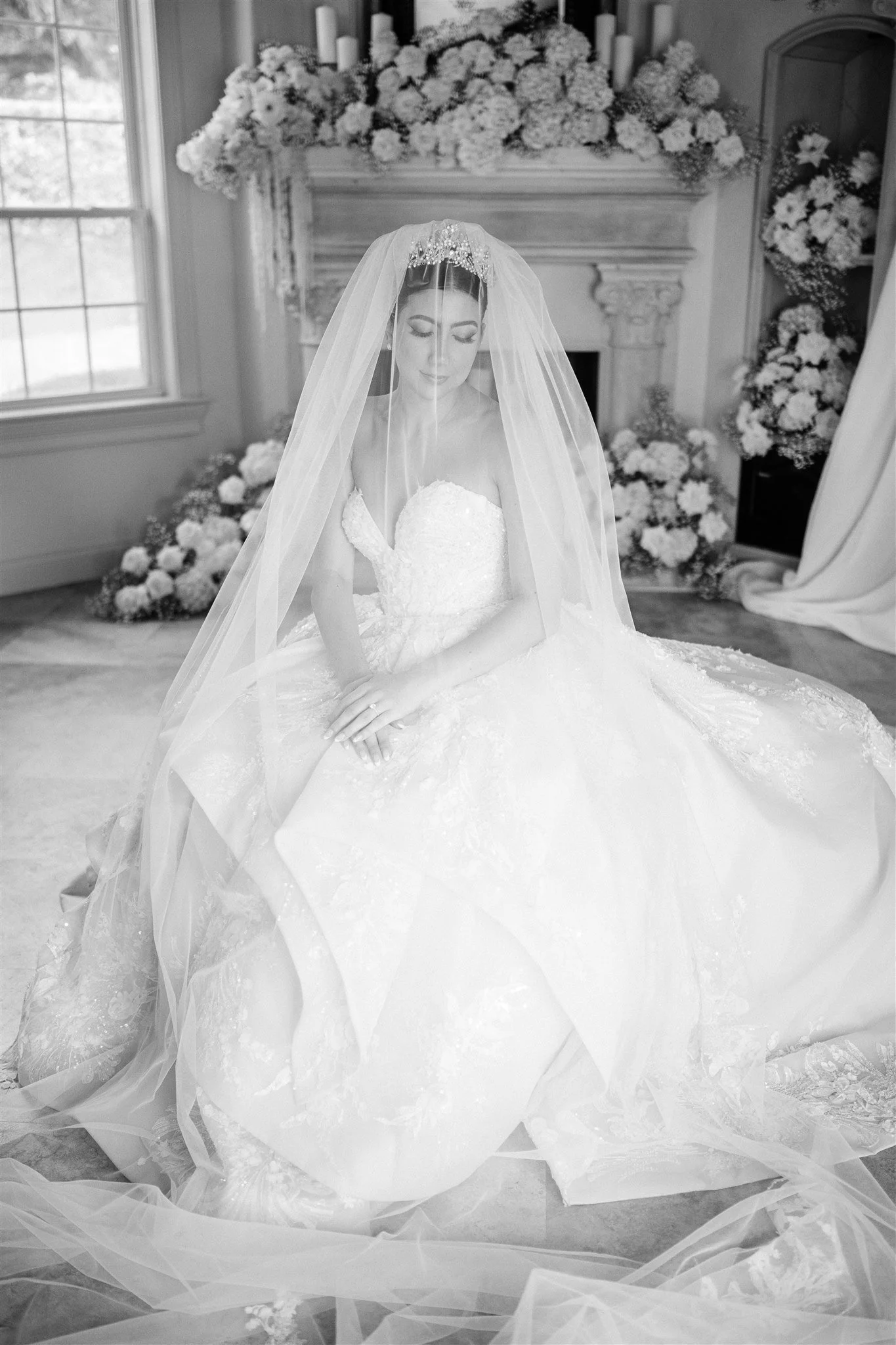 A bride in a wedding gown and veil sitting in front of a fireplace decorated with flowers.
