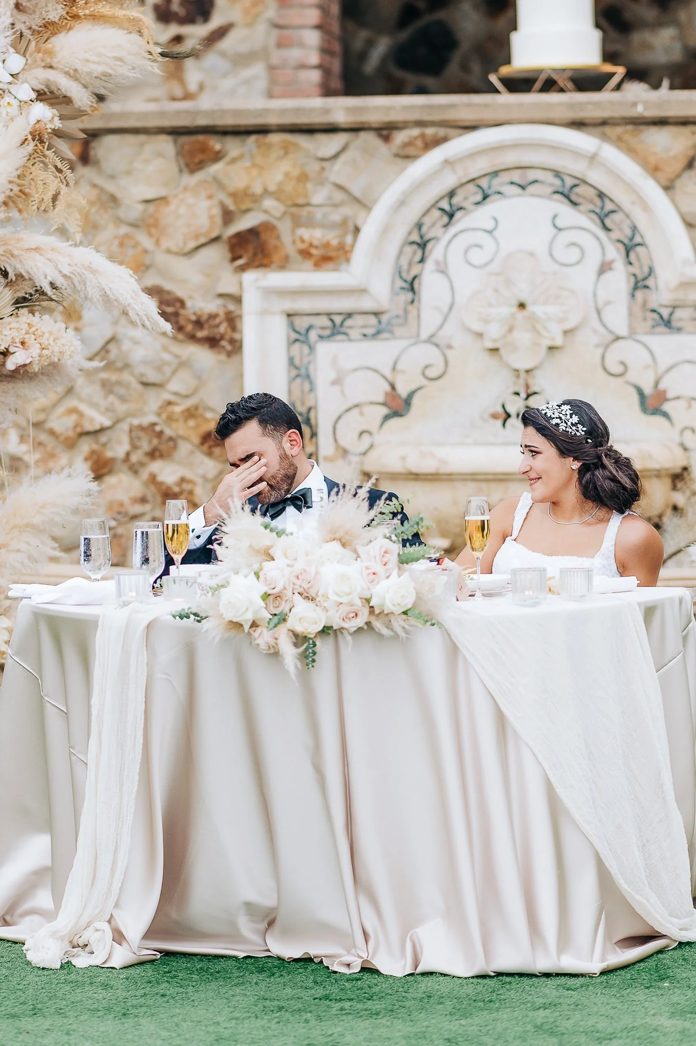 A bride and groom at a wedding reception, sitting at a table decorated with flowers, with the bride smiling and the groom covering his face with his hand.