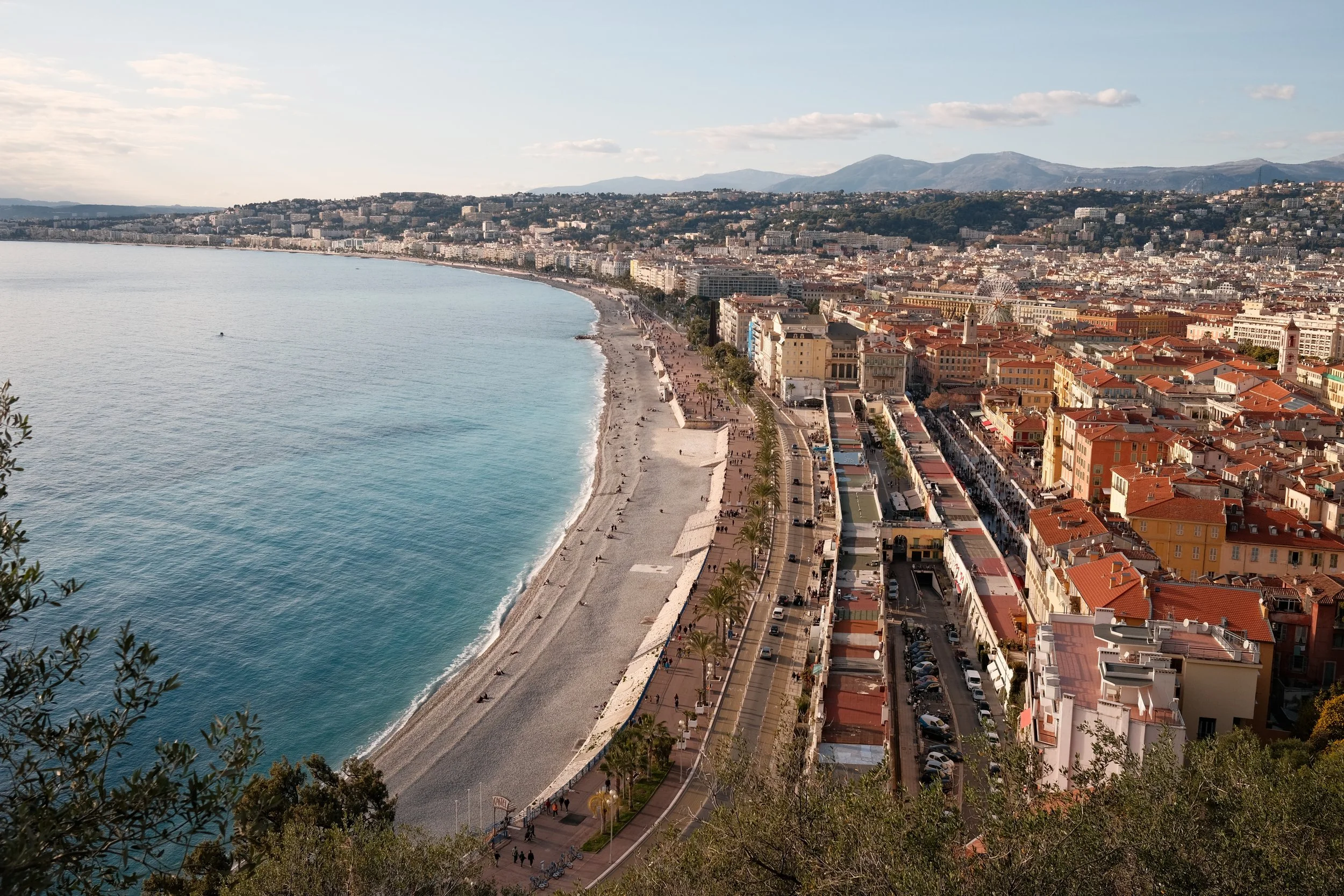 Aerial view of a seaside city with a long sandy beach and a promenade lined with palm trees, surrounded by colorful buildings with red roofs, and distant mountains in the background.