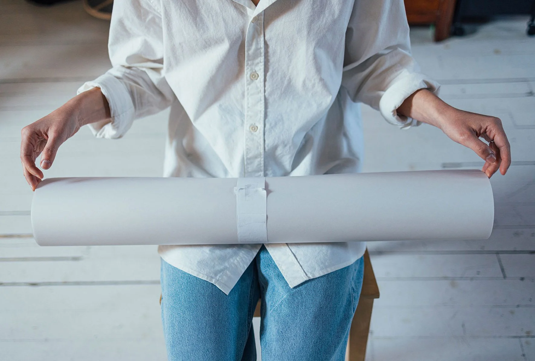 Person in a white shirt and jeans holding a large rolled-up sheet of paper or blueprint.
