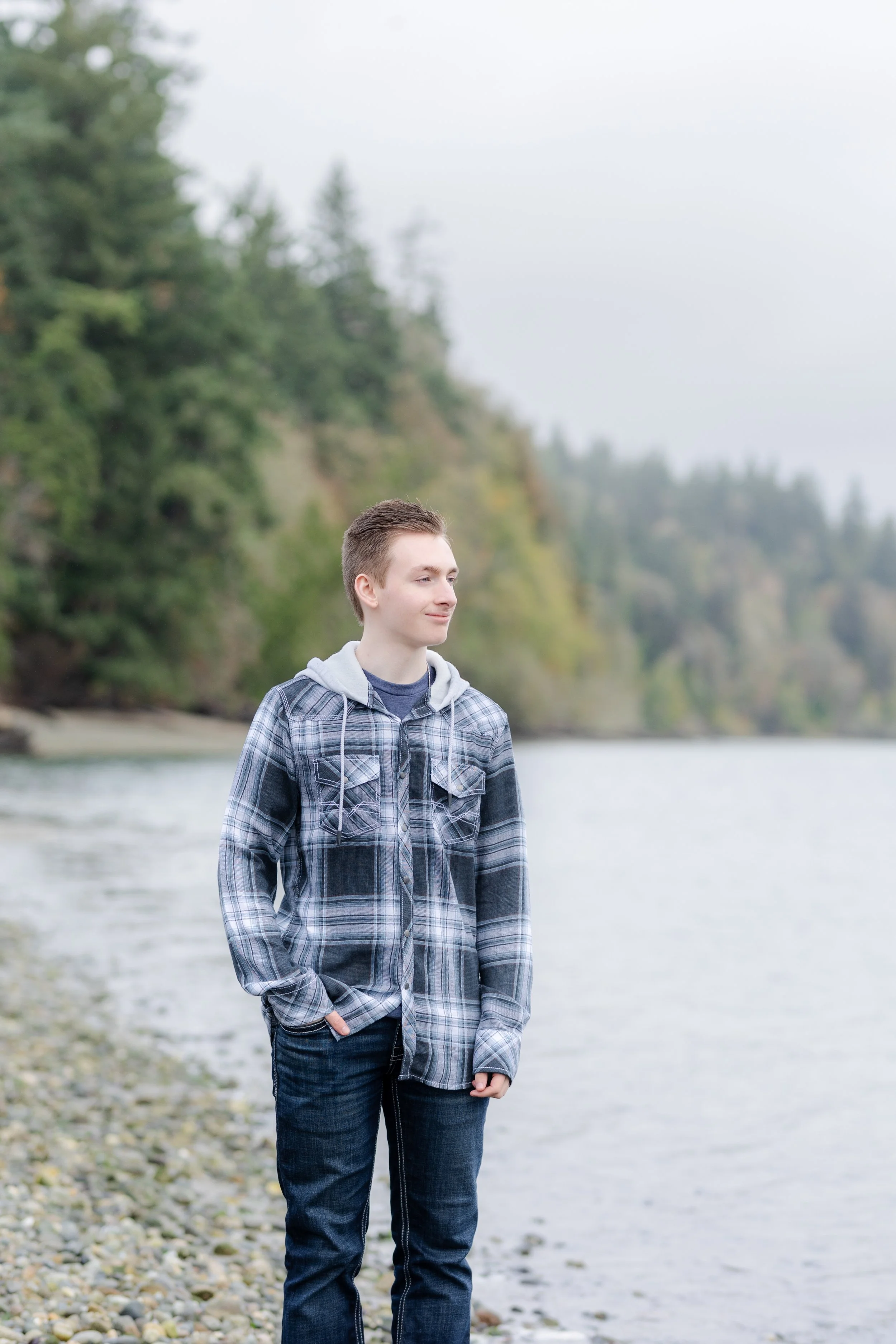 Winter senior photo in Kitsap County, Washington of boy wearing flannel button up jacket standing in front of the water on a beach on a cloudy day.