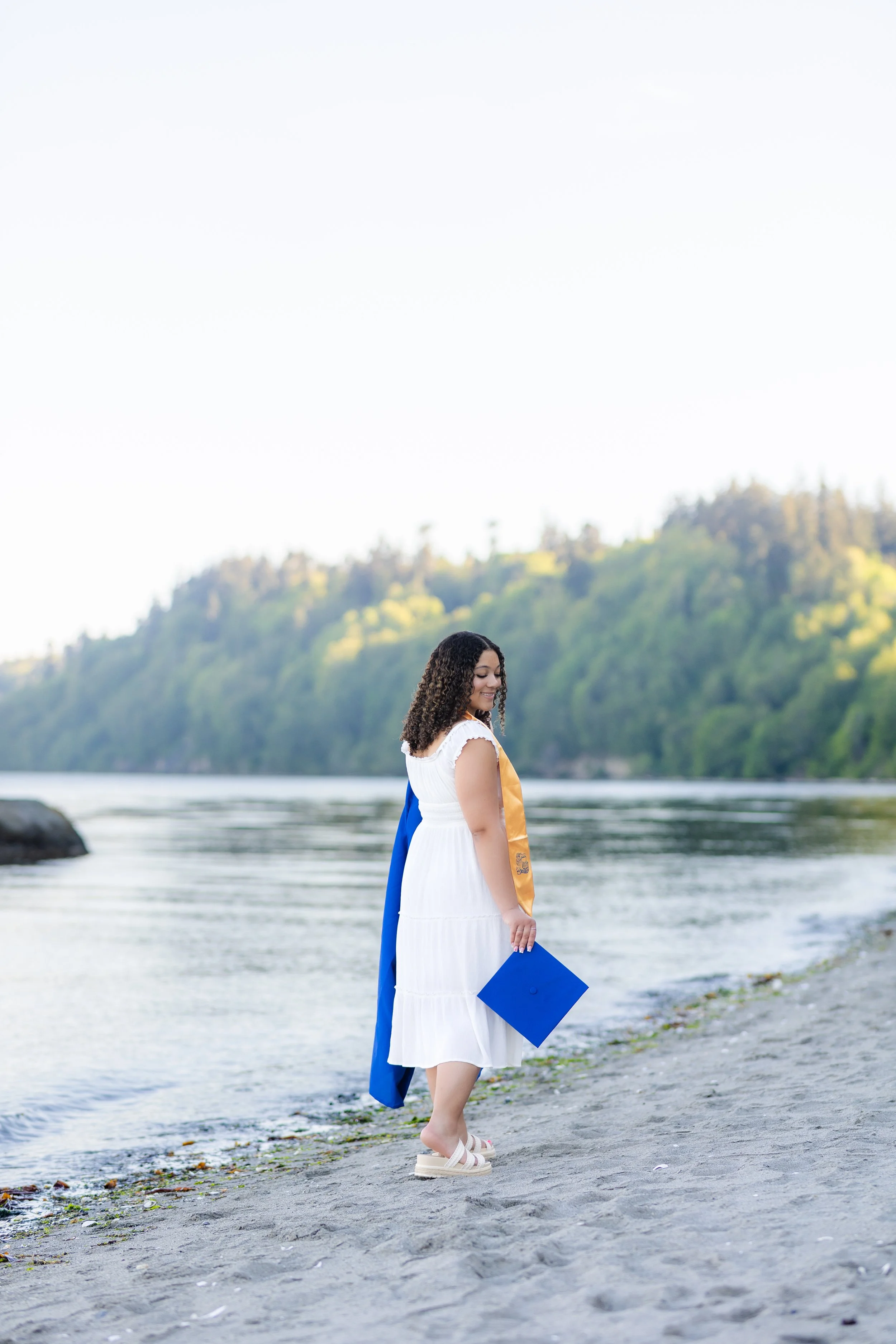 Kitsap graduation picture of a girl walking along the beach holding her cap and gown and wearing her sash