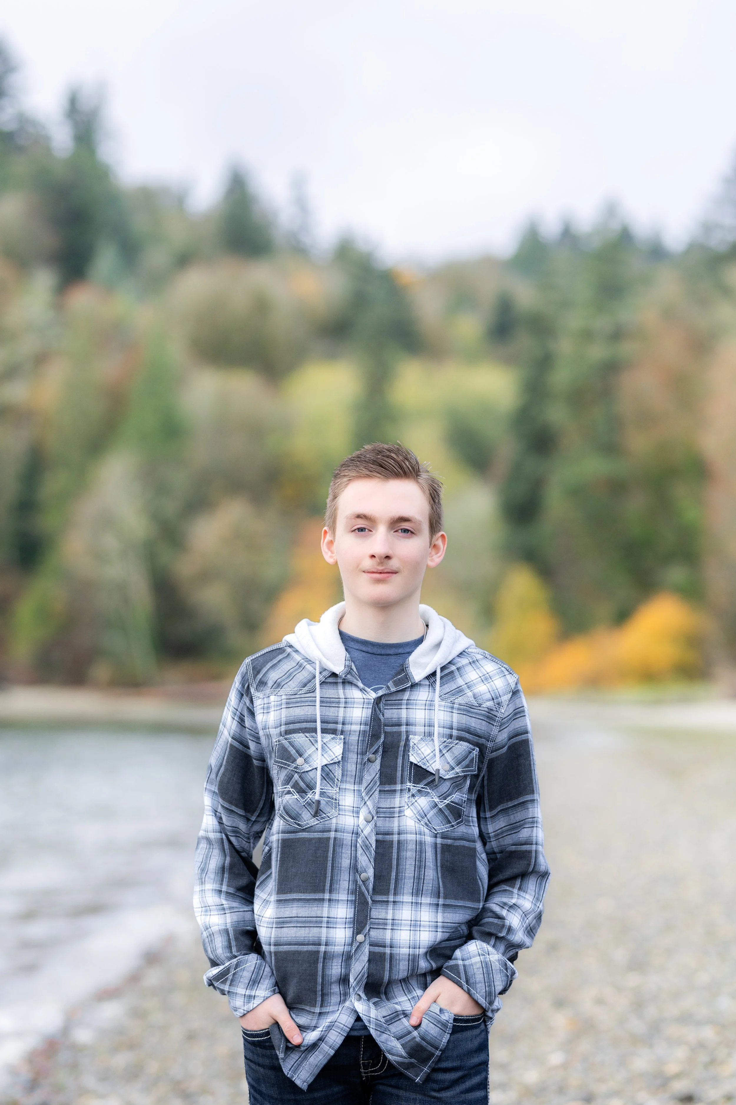 Fall senior picture in Kitsap County, Washington of senior boy looking at camera with fall trees and green trees behind him on a beach.