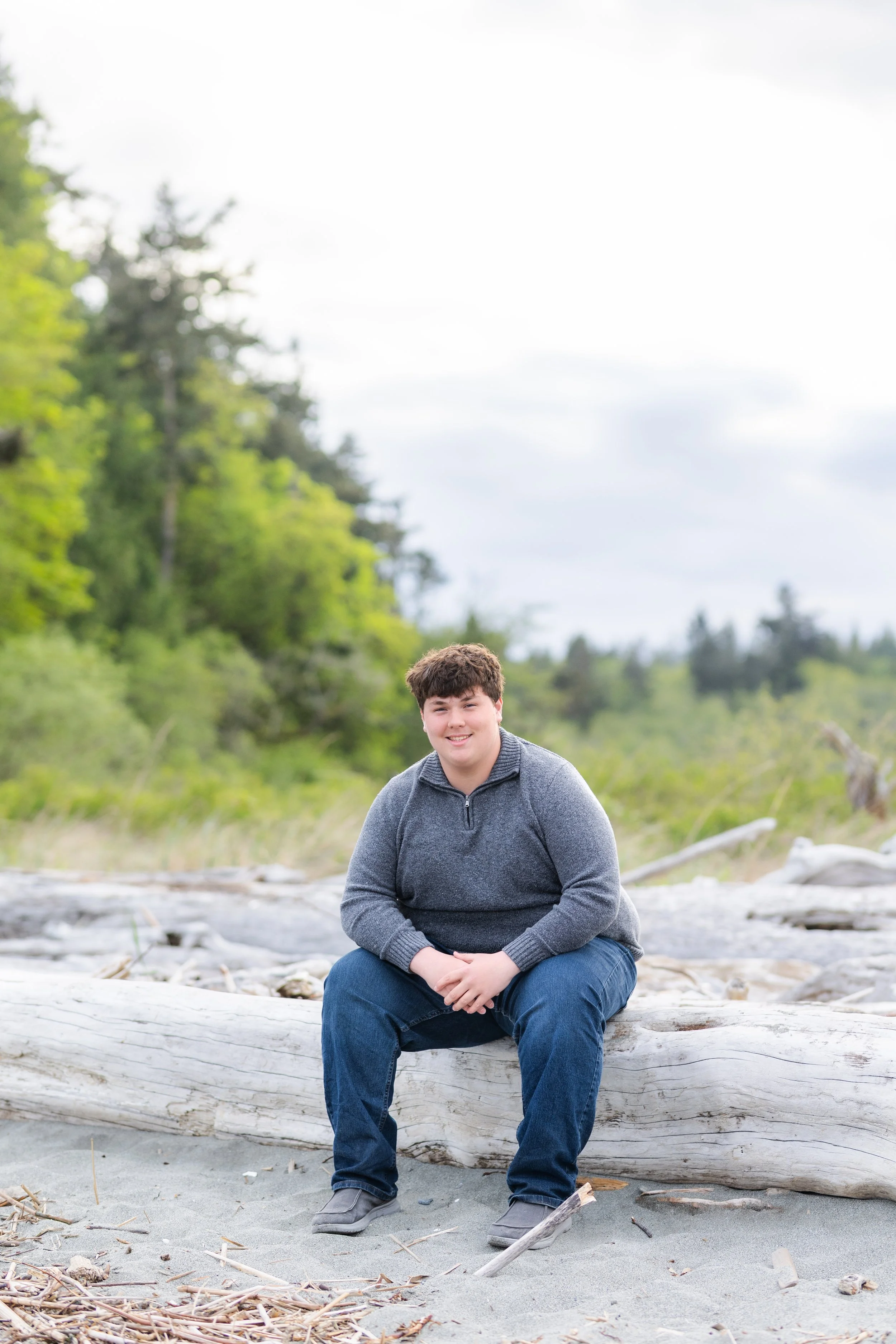 Kitsap senior photo taken of a boy sitting on driftwood with green trees behind him in Kingston, WA