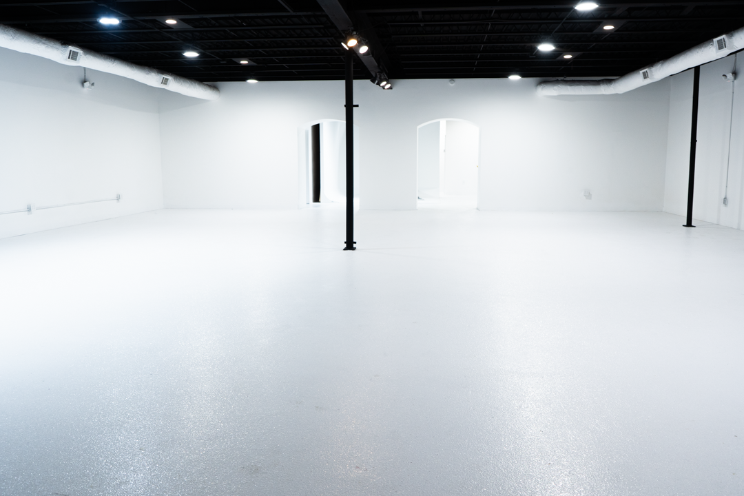 An empty, white-walled room with black ceiling and support beams, bright lighting, and two arched doorways.