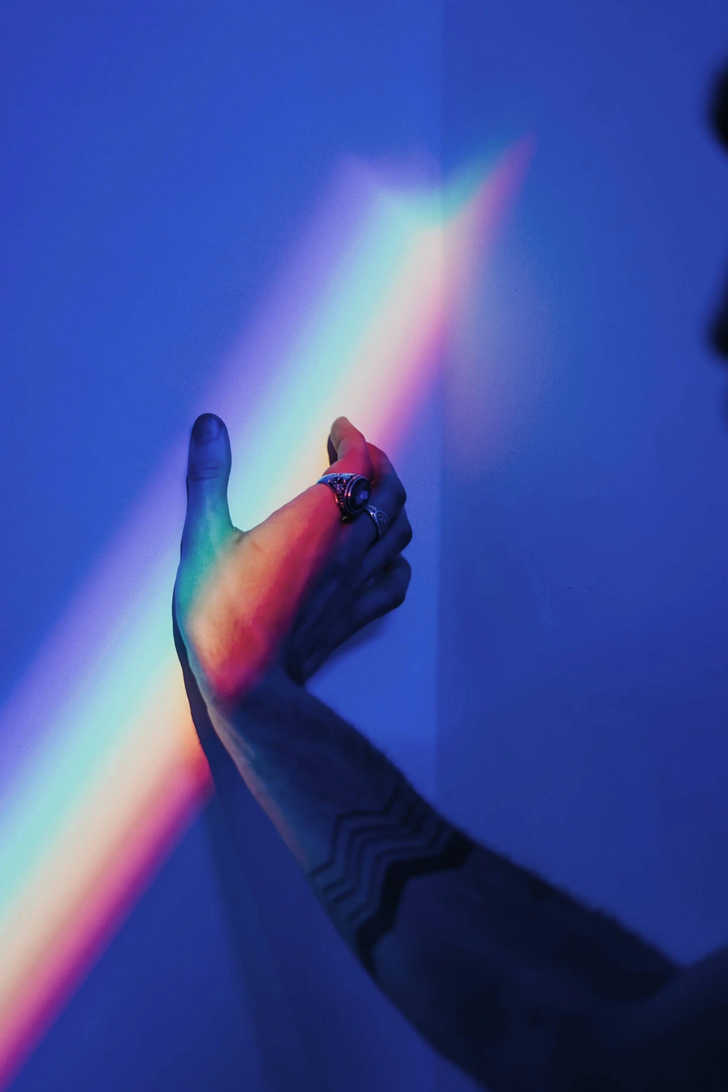 An outstretched hand wearing many rings touching a wall with a band of rainbow light across it.  The arm of the hand has a geometric tattoo