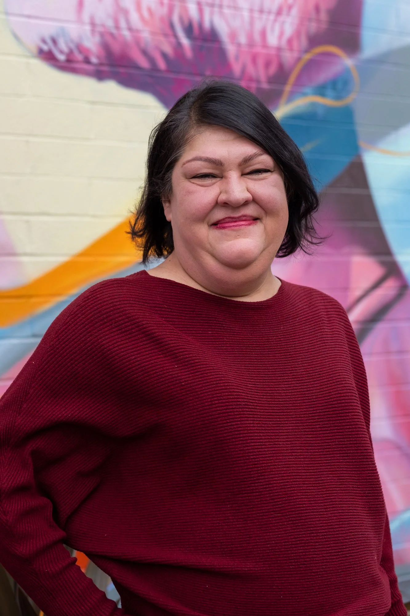 Tall, woman with light tan skin and a chin length dark brown bob wearing a bugandy shirt seen from the waist up standing in front of a colorful mural of flowers and geometric shapes