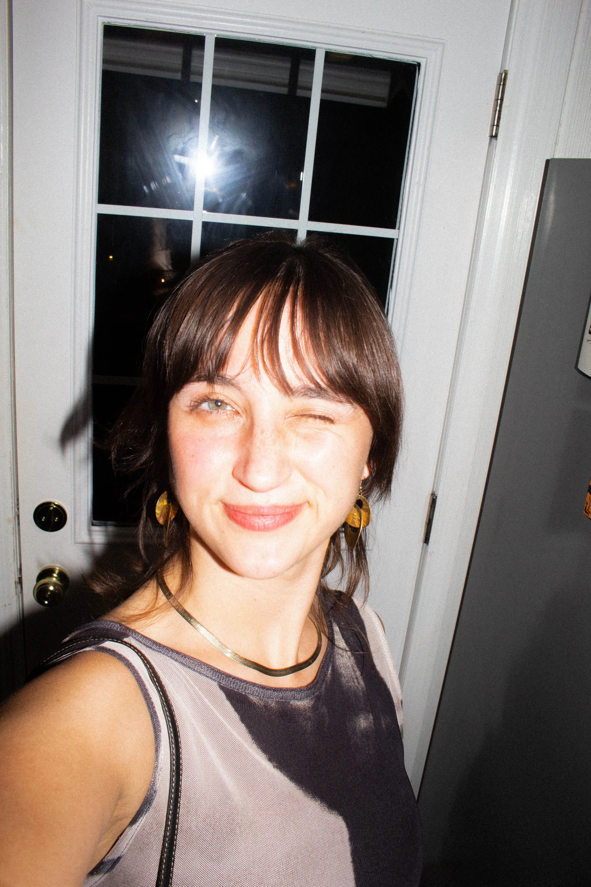 Artist self portrait - A woman with short brown hair, wearing earrings and a sleeveless top, taking a selfie in front of a window at night.