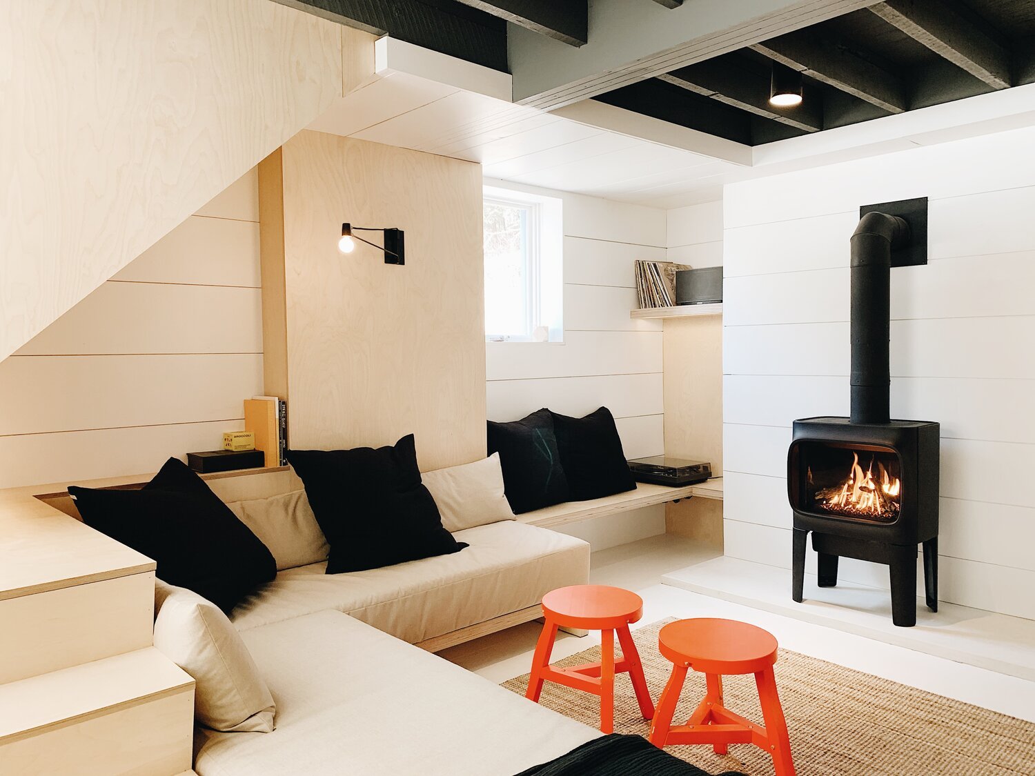 Modern living room with light wood paneling, a beige sectional sofa, black cushions, orange stools, and a wood-burning stove with a visible fire. Exposed ceiling beams and a cozy, minimalist aesthetic.