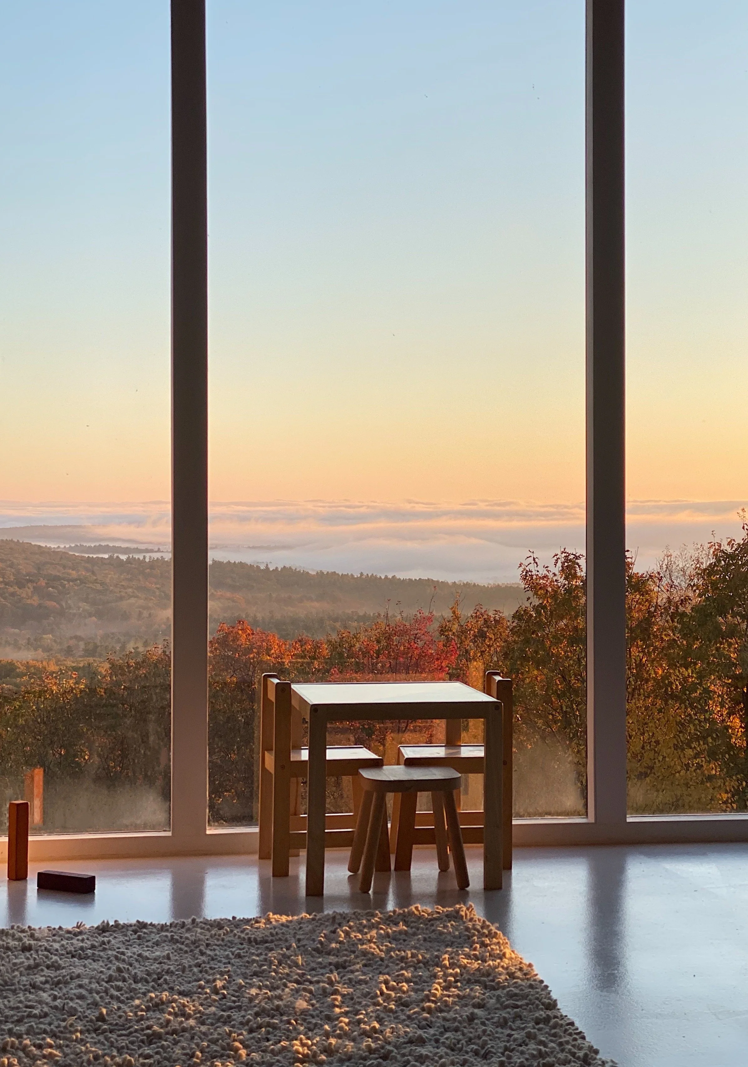 A cozy room with a wooden table and chairs in front of large windows showcasing a scenic view of a forested landscape and sunset sky.