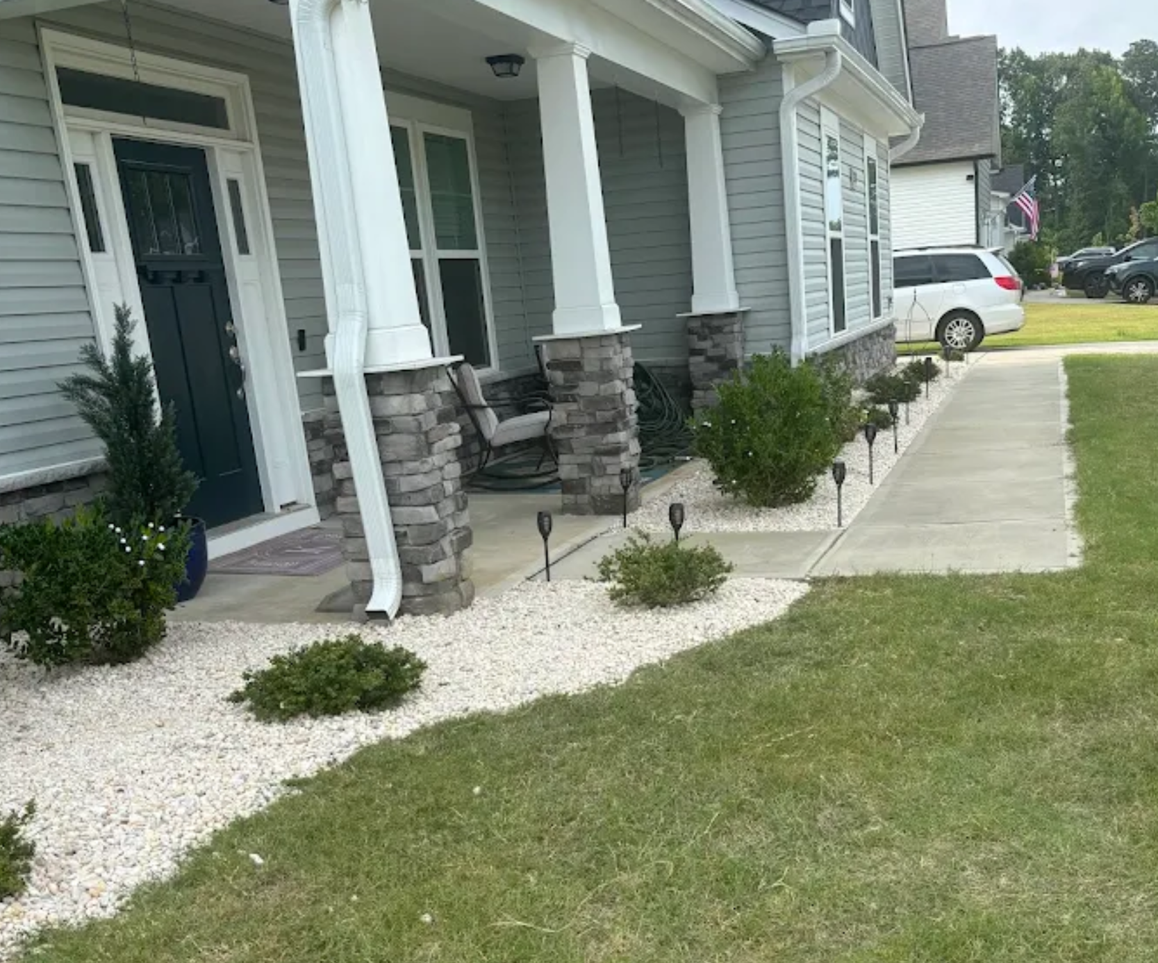 Front porch with stone columns, dark door, paved walkway, white gravel landscaping, green shrubs, and an SUV parked in the driveway.