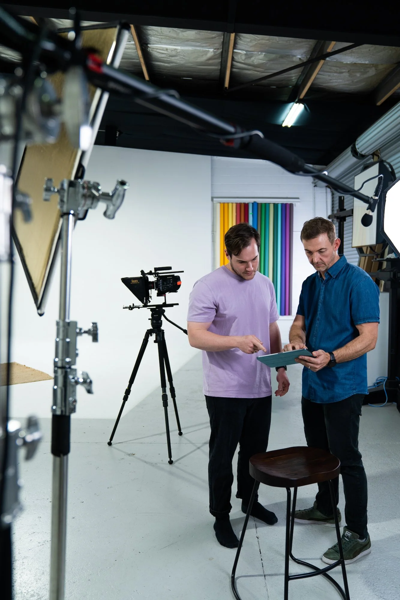 Two men in a photo studio, one holding a tablet, with a professional camera on a tripod and colorful backgrounds.