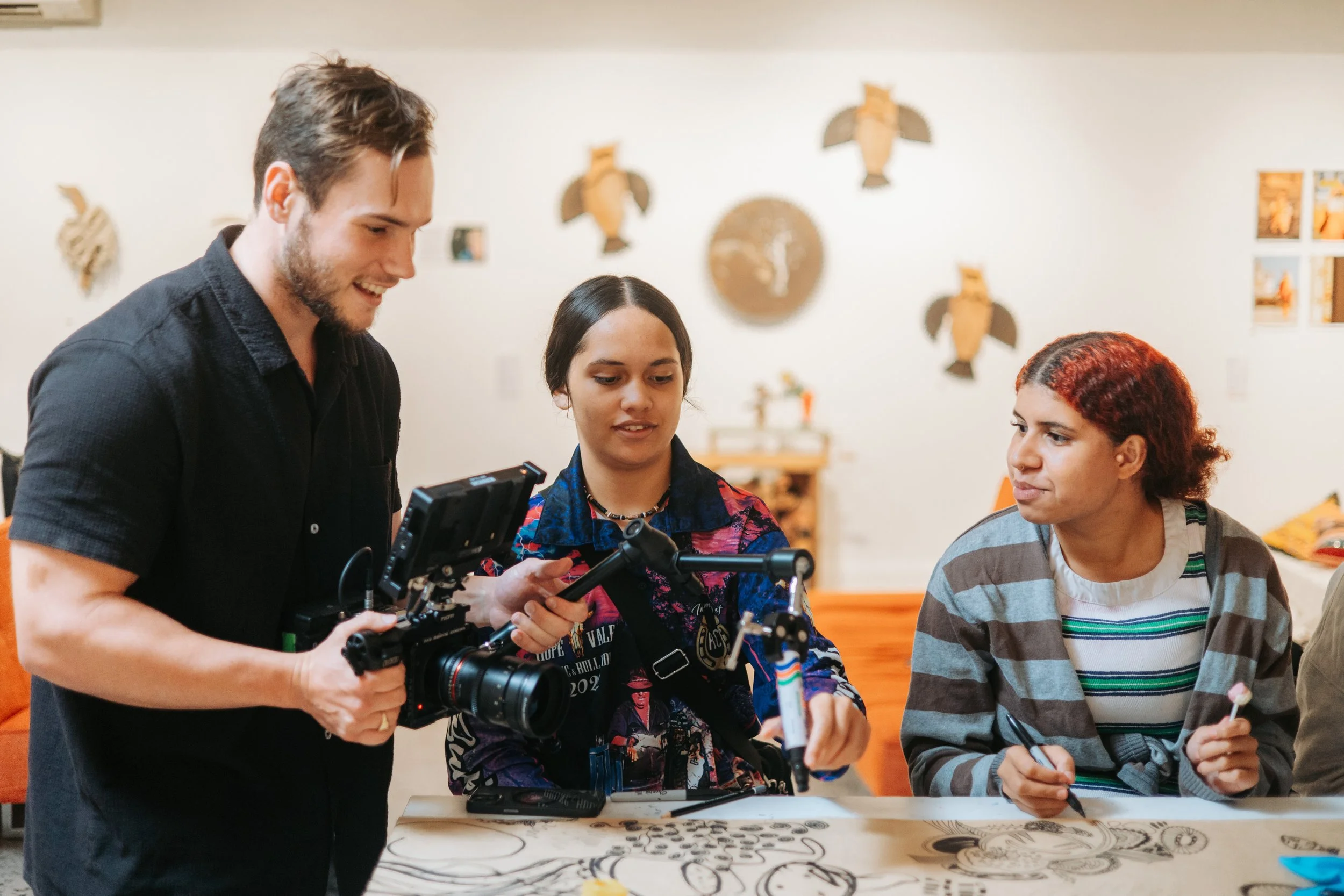 Three people engaging in an art activity, one filming with a camera and two working on a drawing at a table, in an art studio setting.