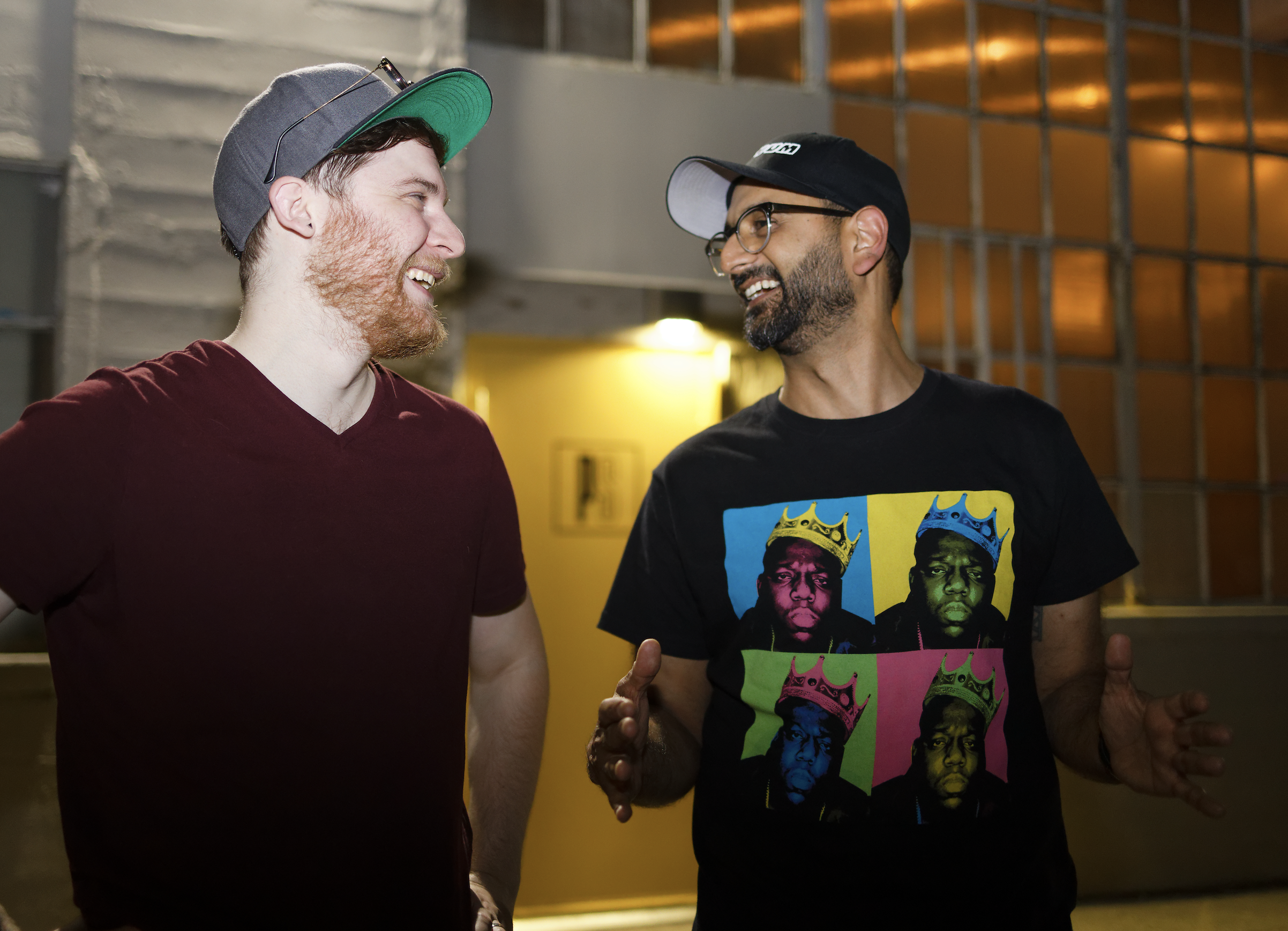 Two men are smiling and talking to each other, one wearing a dark red V-neck shirt and a gray cap, and the other wearing a black T-shirt with colorful pop art and a black cap in an indoor setting with warm lighting.