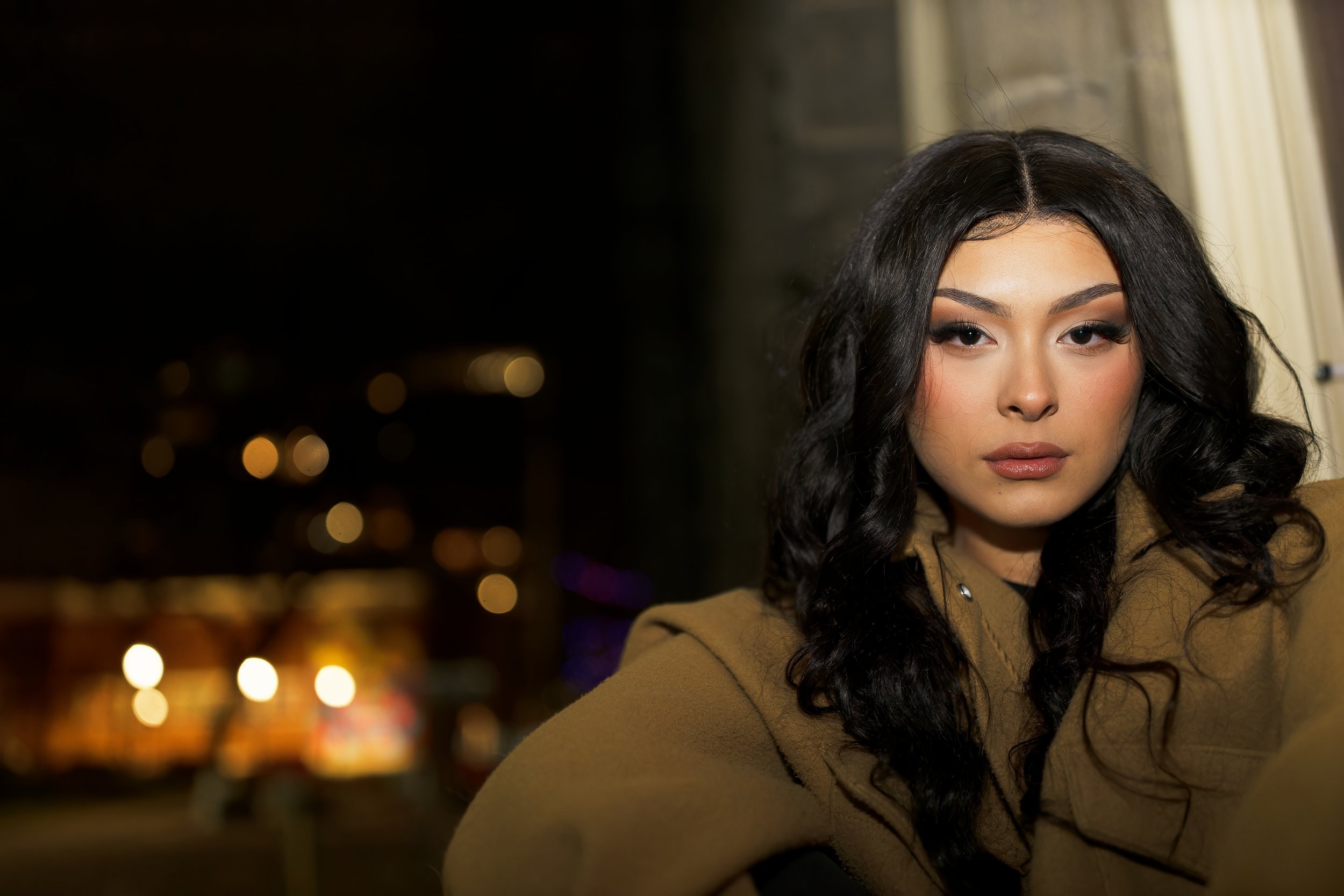 A young woman with dark, wavy hair sitting indoors near a window with blurred city lights in the background.