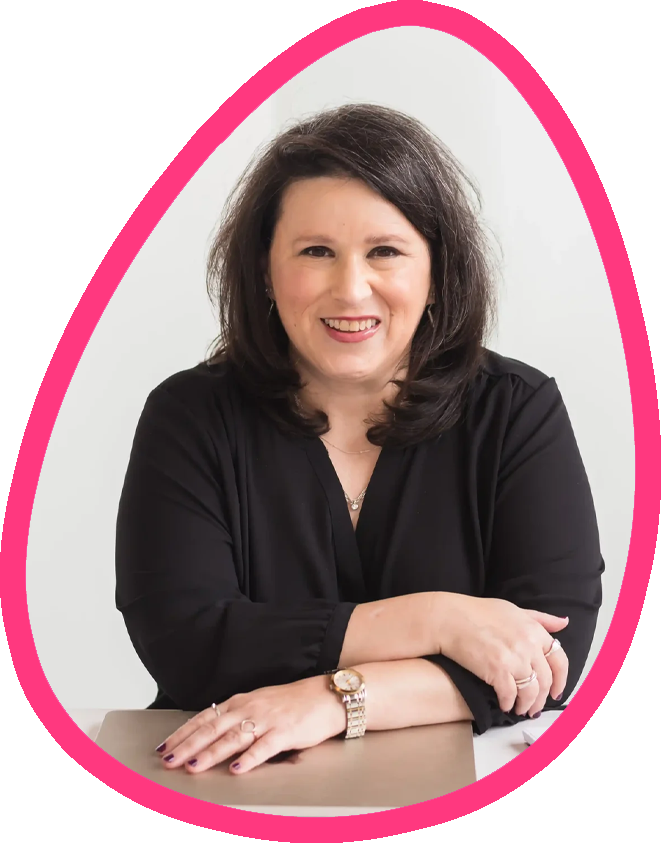 Carrie Lubing smiling, wearing a black top, gold watch, and jewelry, sitting at a table with arms crossed, framed by a pink and white background.