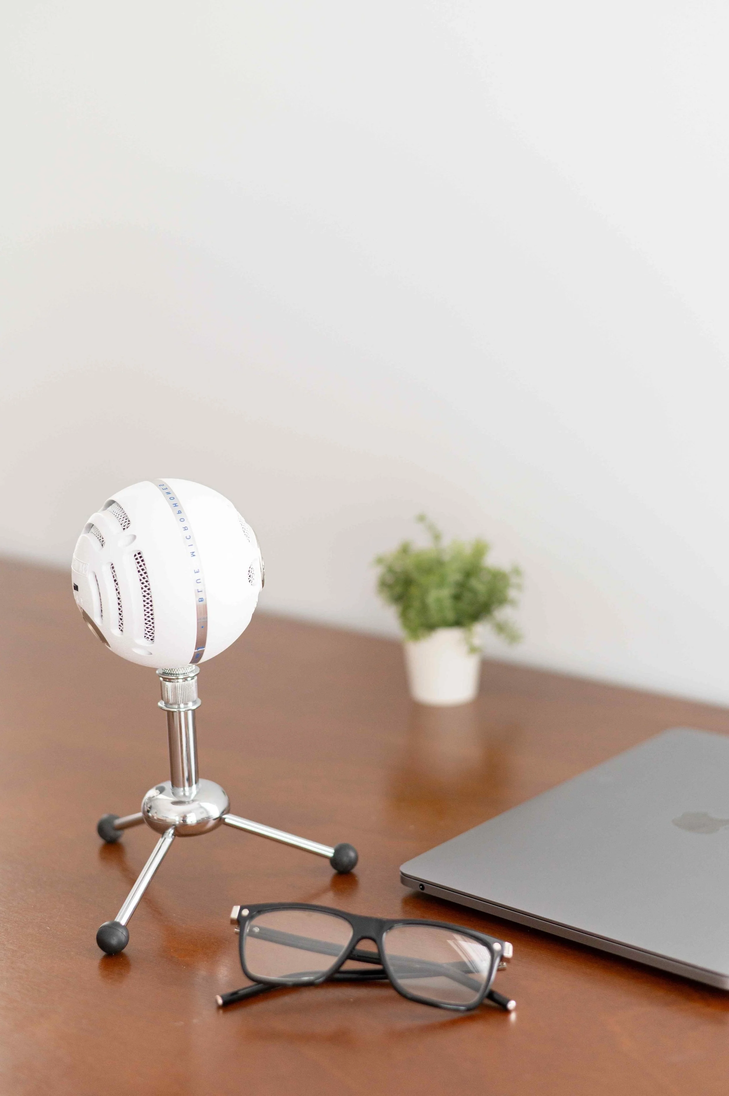 A workspace with a gray laptop, black glasses, a small white potted plant, and a white microphone on a wooden desk.