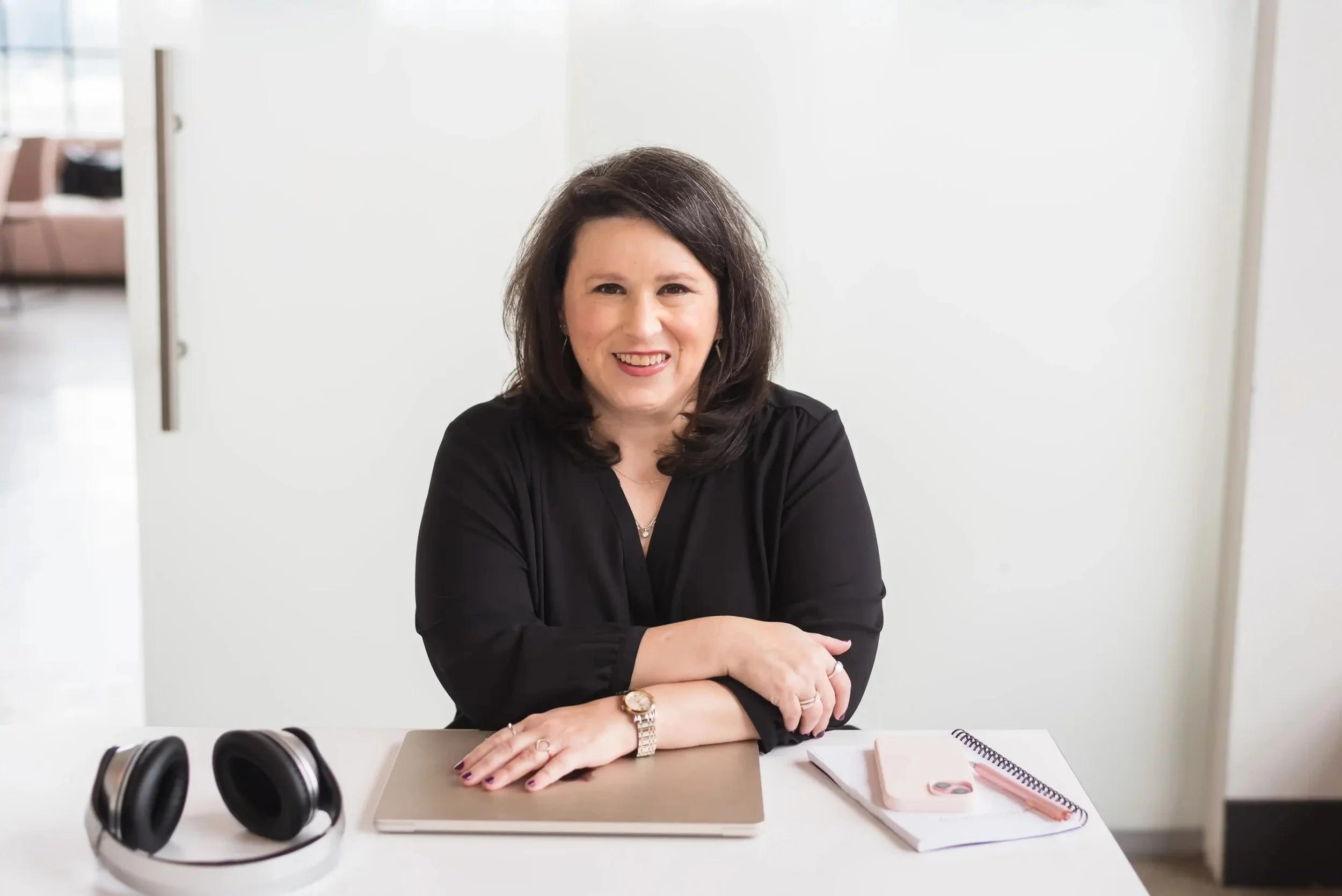 Carrie sitting at a white desk, smiling, with headphones, a laptop, a notebook, and a phone on the desk.