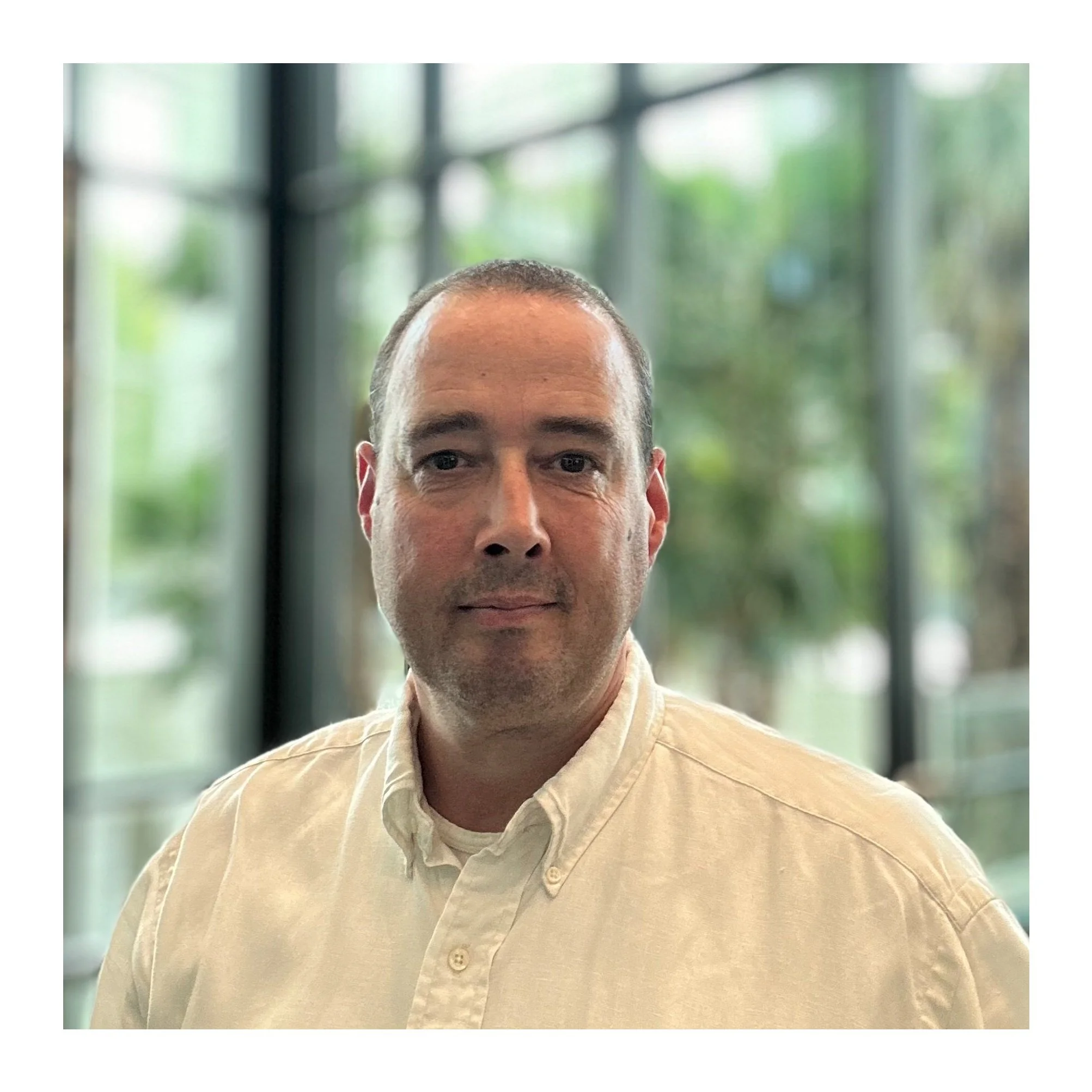 Portrait of Maarten with short brown hair, wearing a white collared shirt, standing indoors with large windows and greenery in the background.