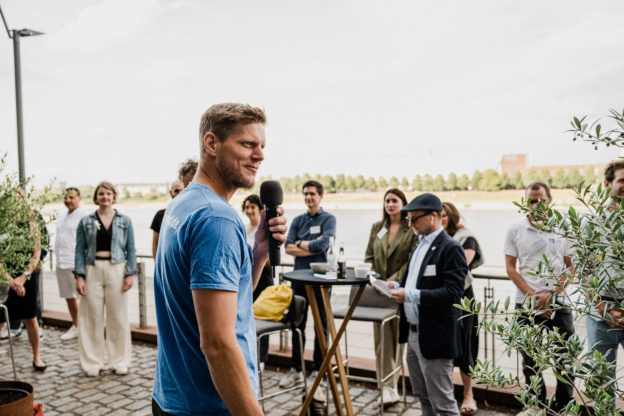 Mann in blauer T-Shirt hält ein Mikrofon bei einer outdoor Veranstaltung, umgeben von mehreren Menschen, einige mit Namensschildern, vor Wasser mit Ufer und Bäumen im Hintergrund.