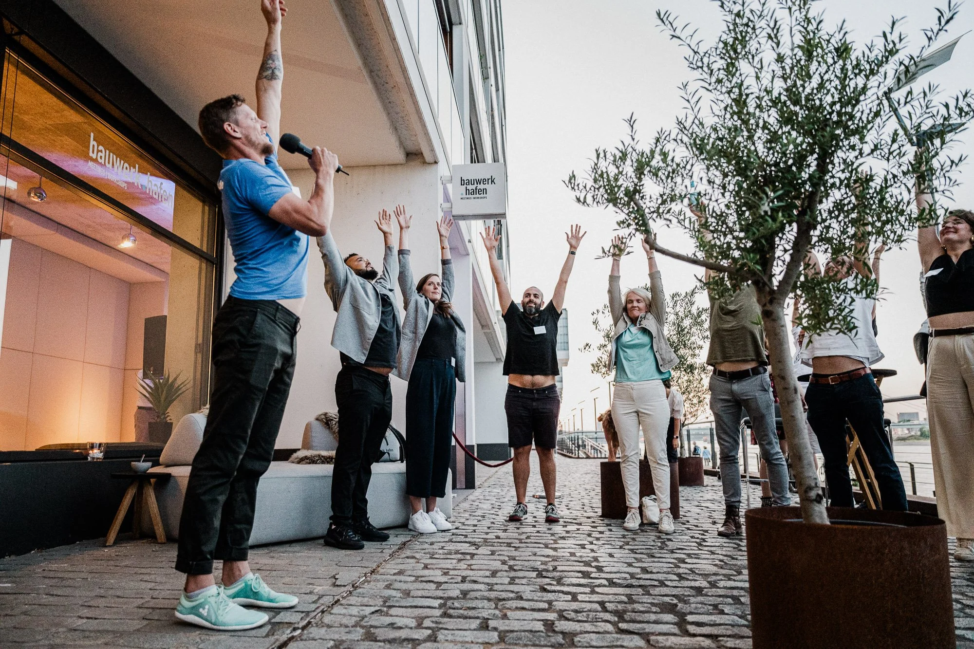 Menschen stehen auf einer Steinstraße außerhalb eines Gebäudes, der Mann in der blauen T-Shirt spricht in ein Mikrofon und die Gruppe hebt die Arme in die Luft.
