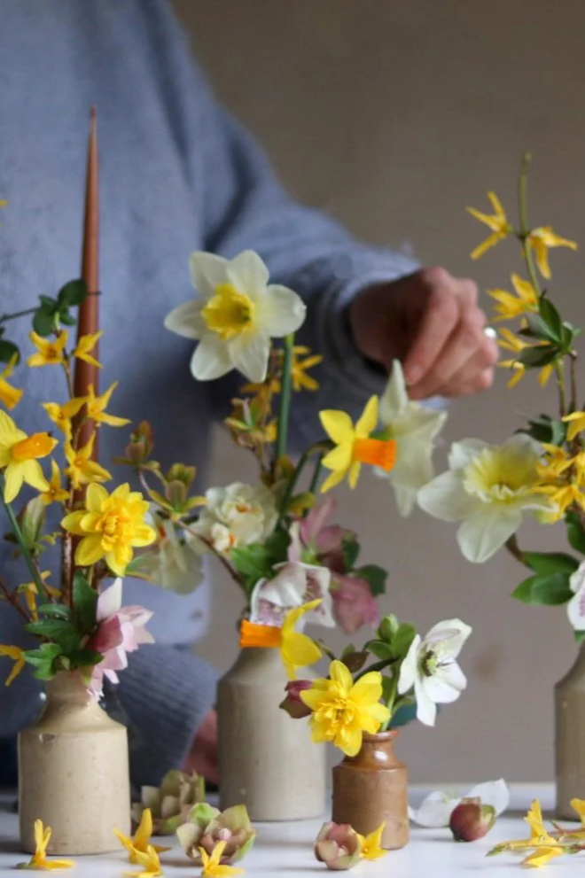 Person arranging flowers in small vases with yellow, white, pink, and purple blossoms on a white table.